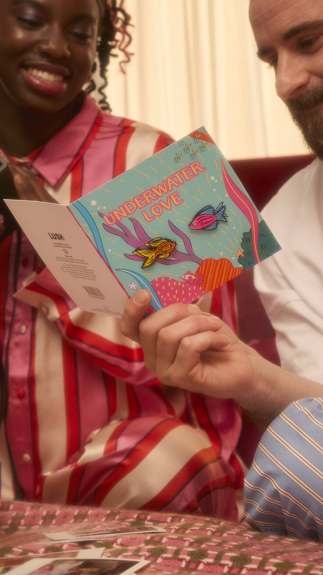 A person holds the open Underwater Love card, revealing the blue cover with pink text and two attached fish pins. Beside them, a smiling companion in pink striped pajamas looks on joyfully.