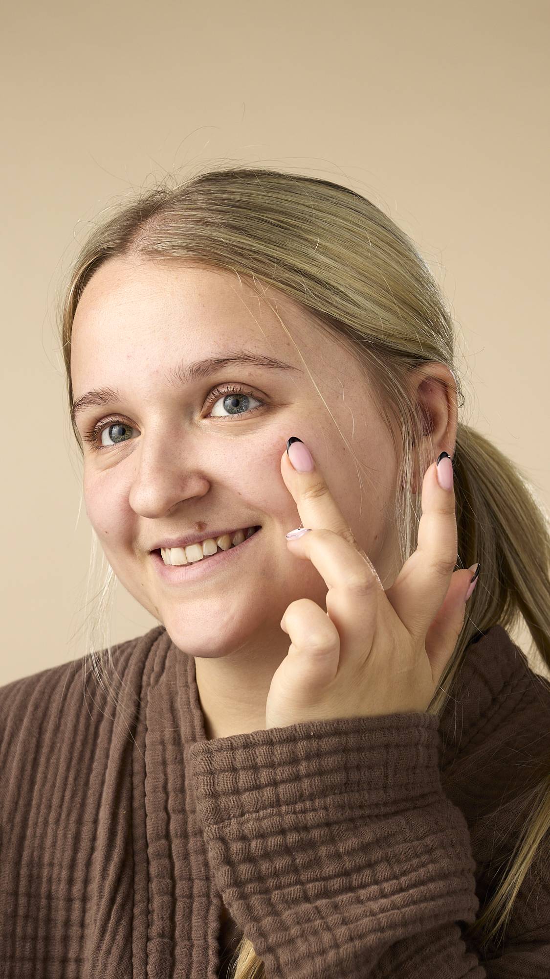 The image shows a light-skinned model with blonde hair pulled back, smiling while applying the Vision facial serum to the cheek under their right eye. They're wearing a brown, textured robe on a light beige background.