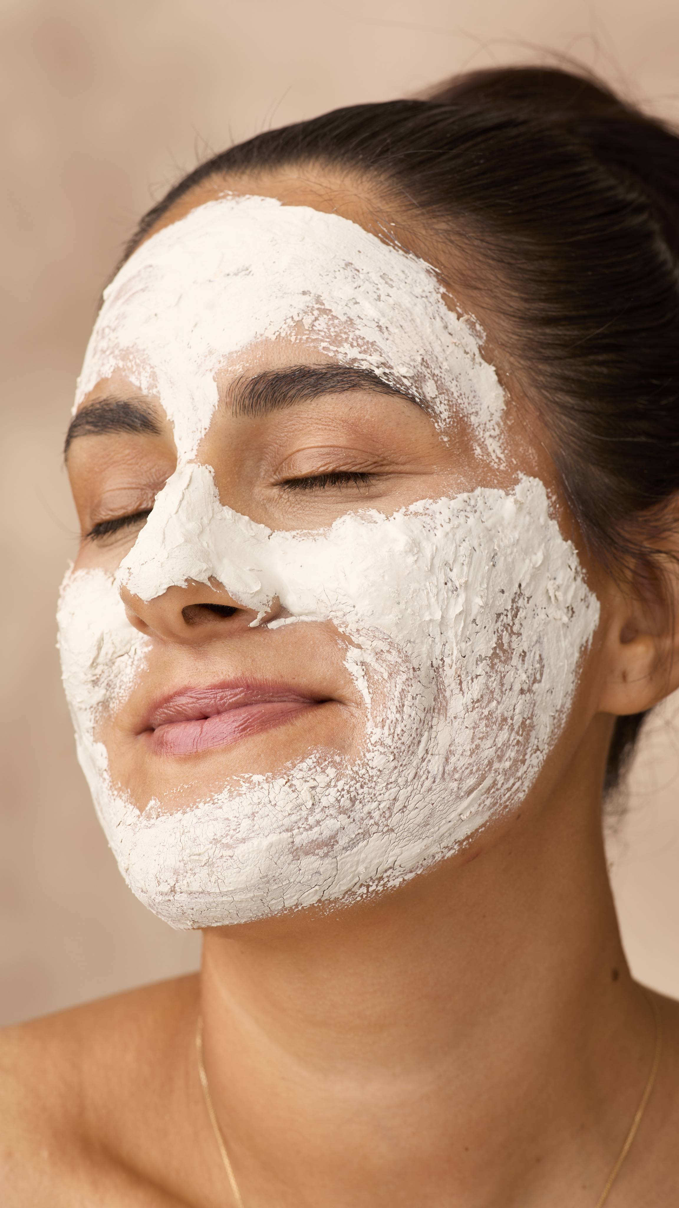 A close-up of a smiling woman with dark hair and eyes closed, relaxed while wearing a thick layer of Lush Who Runs The World? Ghouls! face mask. The textured, light-coloured paste covers her forehead, nose, cheeks and jawline.