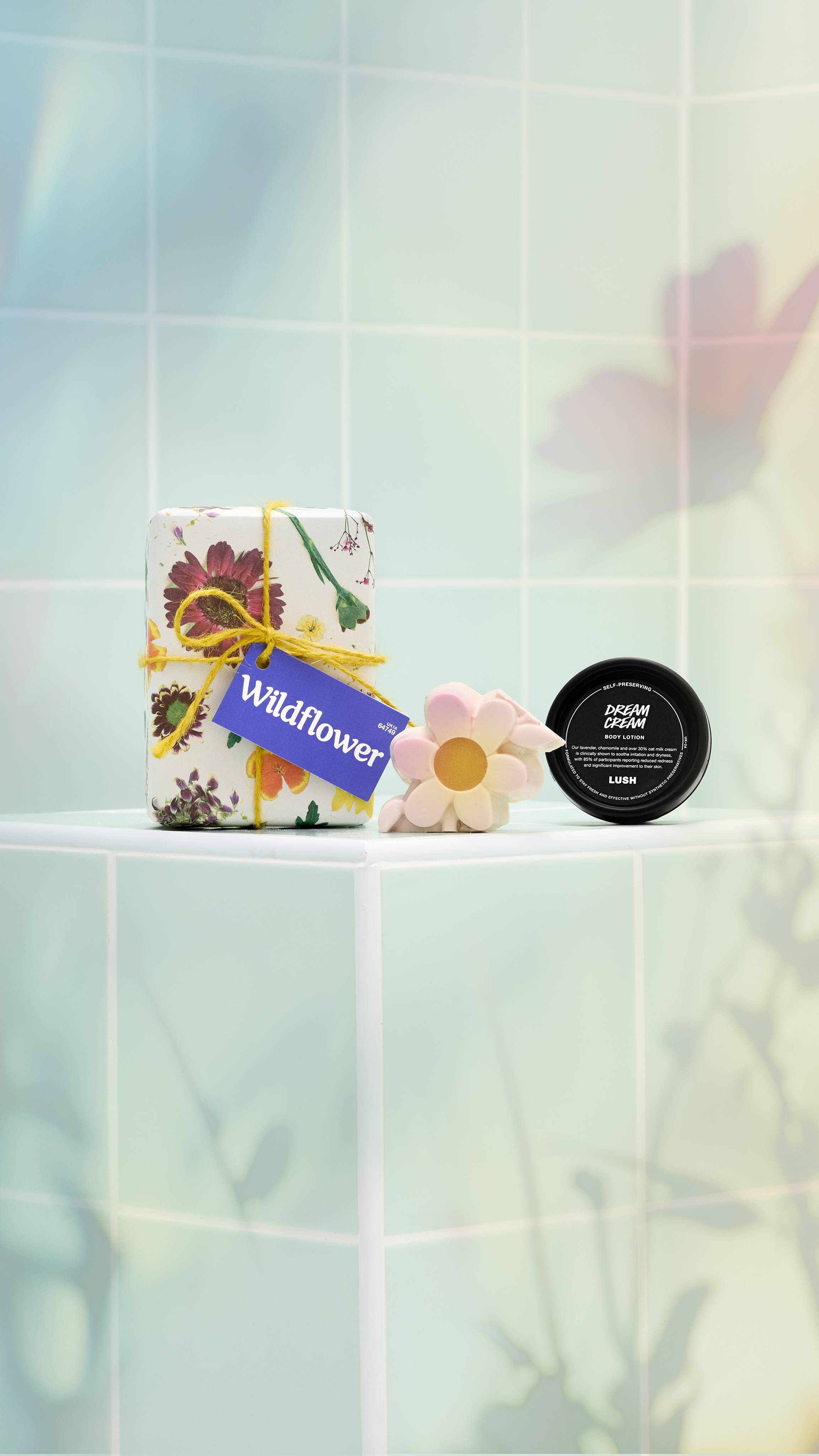 The image shows the Lush "Wildflower" gift set on a light, tiled background. It includes a floral-wrapped box tied with yellow twine and a purple tag, a black pot of "Dream Cream" body lotion, and a pink and yellow flower-shaped soap.