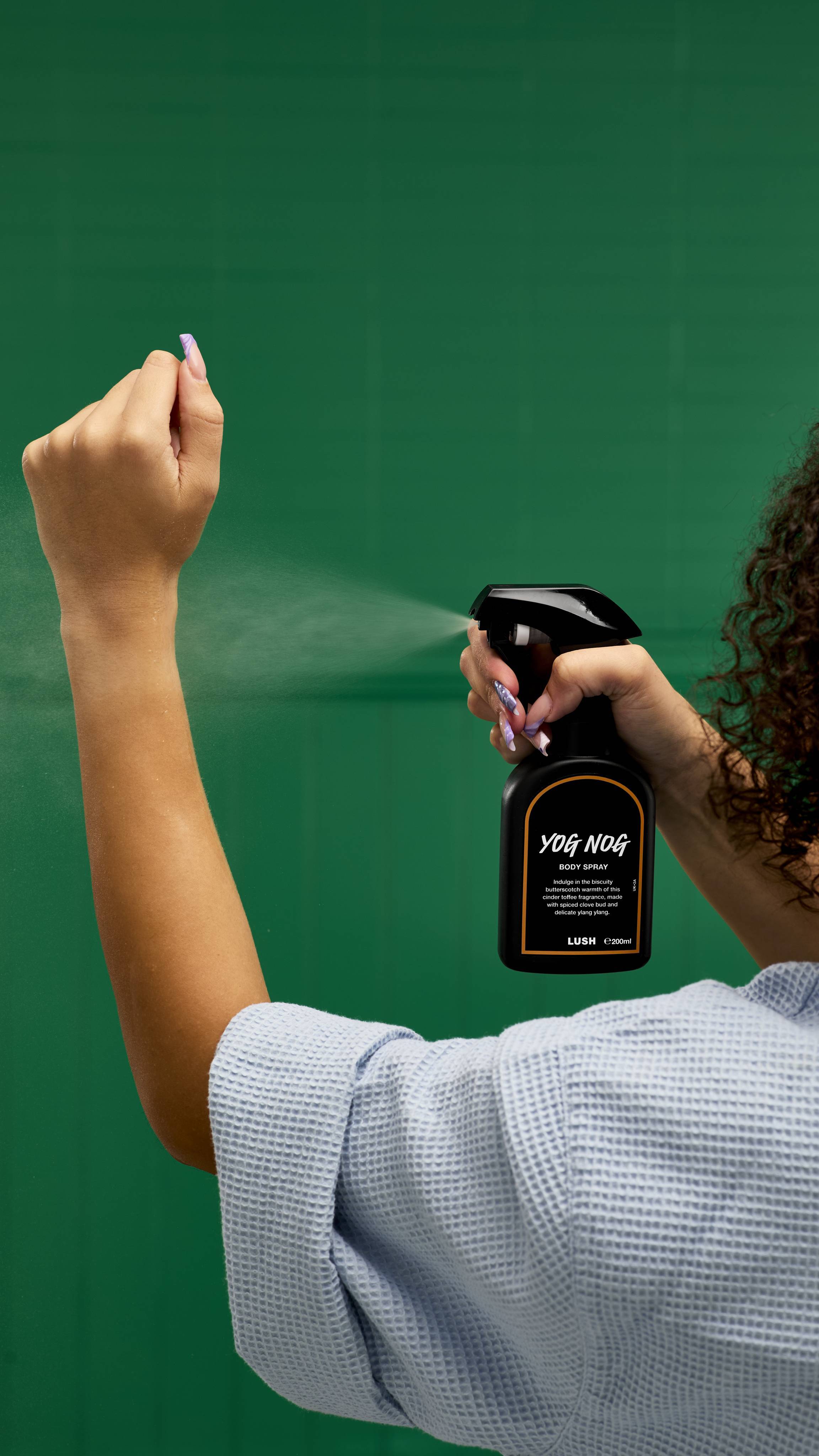 A recycled black LUSH body spray bottle with a trigger nozzle is held by a person from behind. The bottle, with a gold-rimmed "Yog Nog Body Spray" label, is spritzed, and the mist is visible against a vibrant green background.