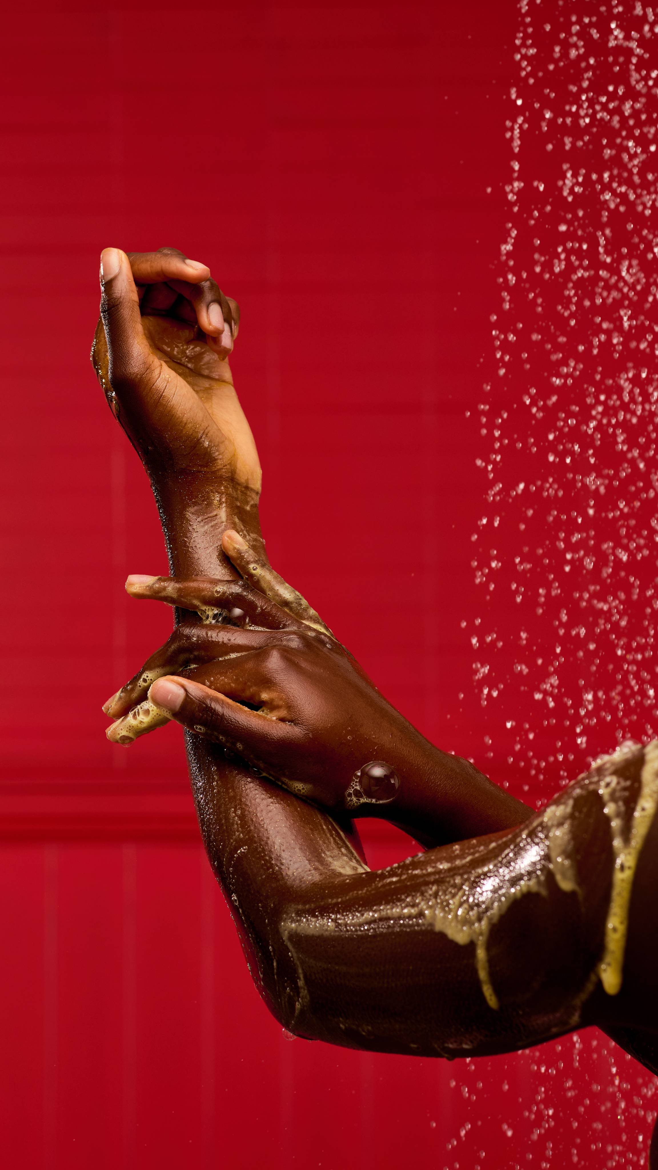 A pair of hands lathers up a glistening, golden-hued product, using LUSH Yog Nog shower gel. Bubbles and a foamy residue are visible on the arms, with water droplets sparkling against a vibrant red background.