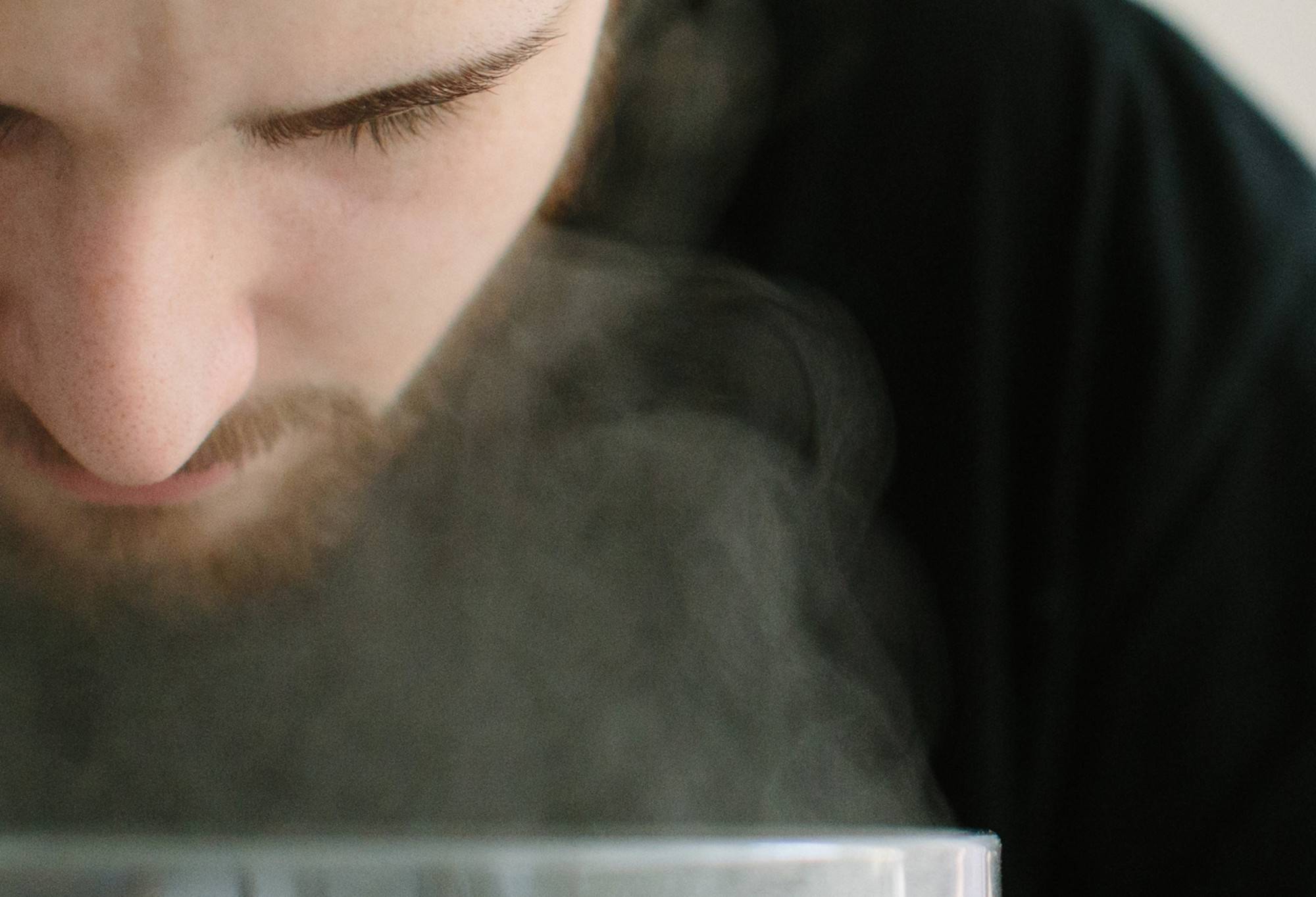 A bearded person hovers their face above a steaming bowl, looking focused yet relaxed.