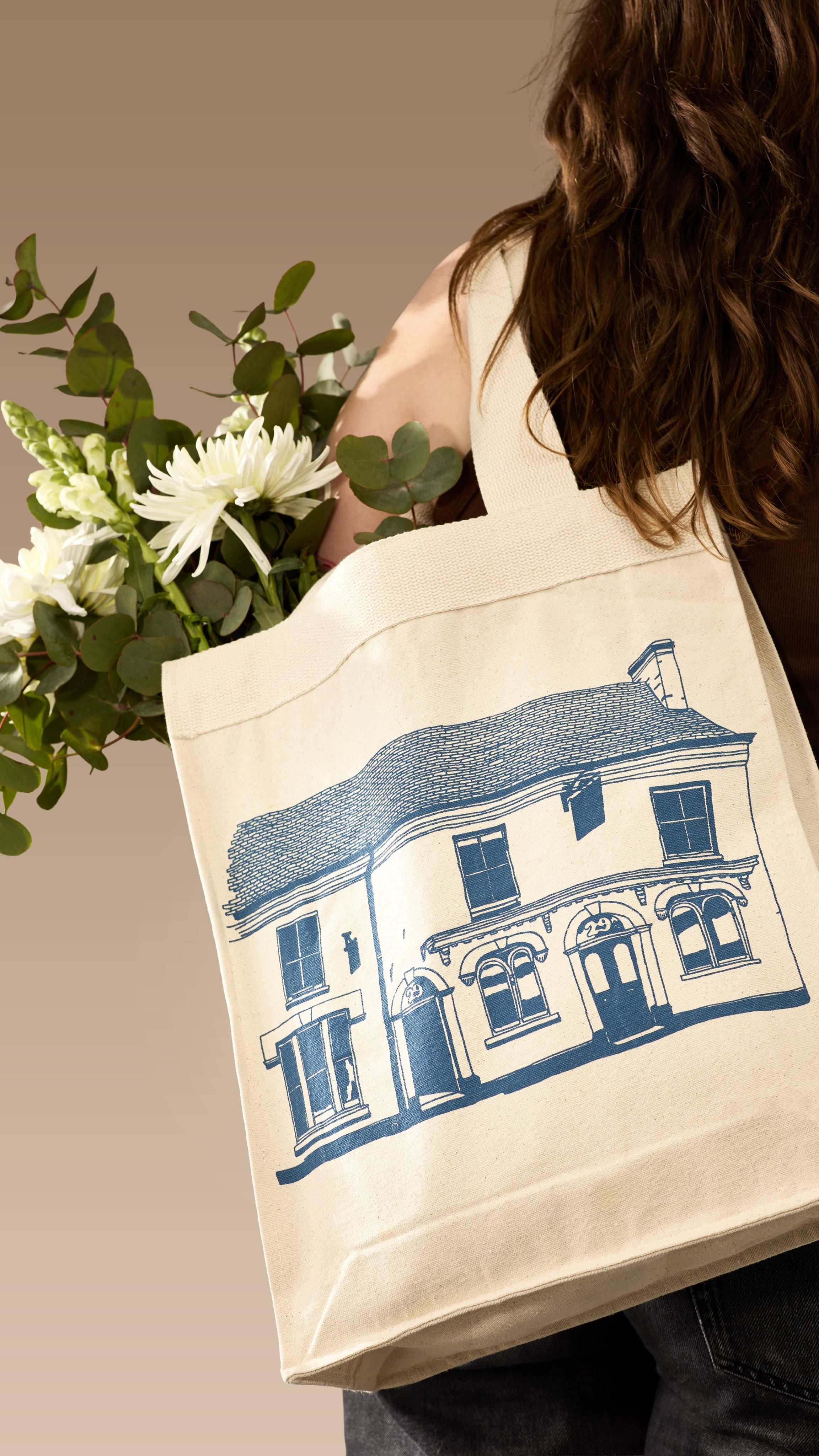 A model with wavy brown hair carries a cream canvas 29 High Street tote bag over their shoulder. The bag, which contains white flowers and eucalyptus, features a navy-blue line drawing of a shop facade, set against a neutral beige background.