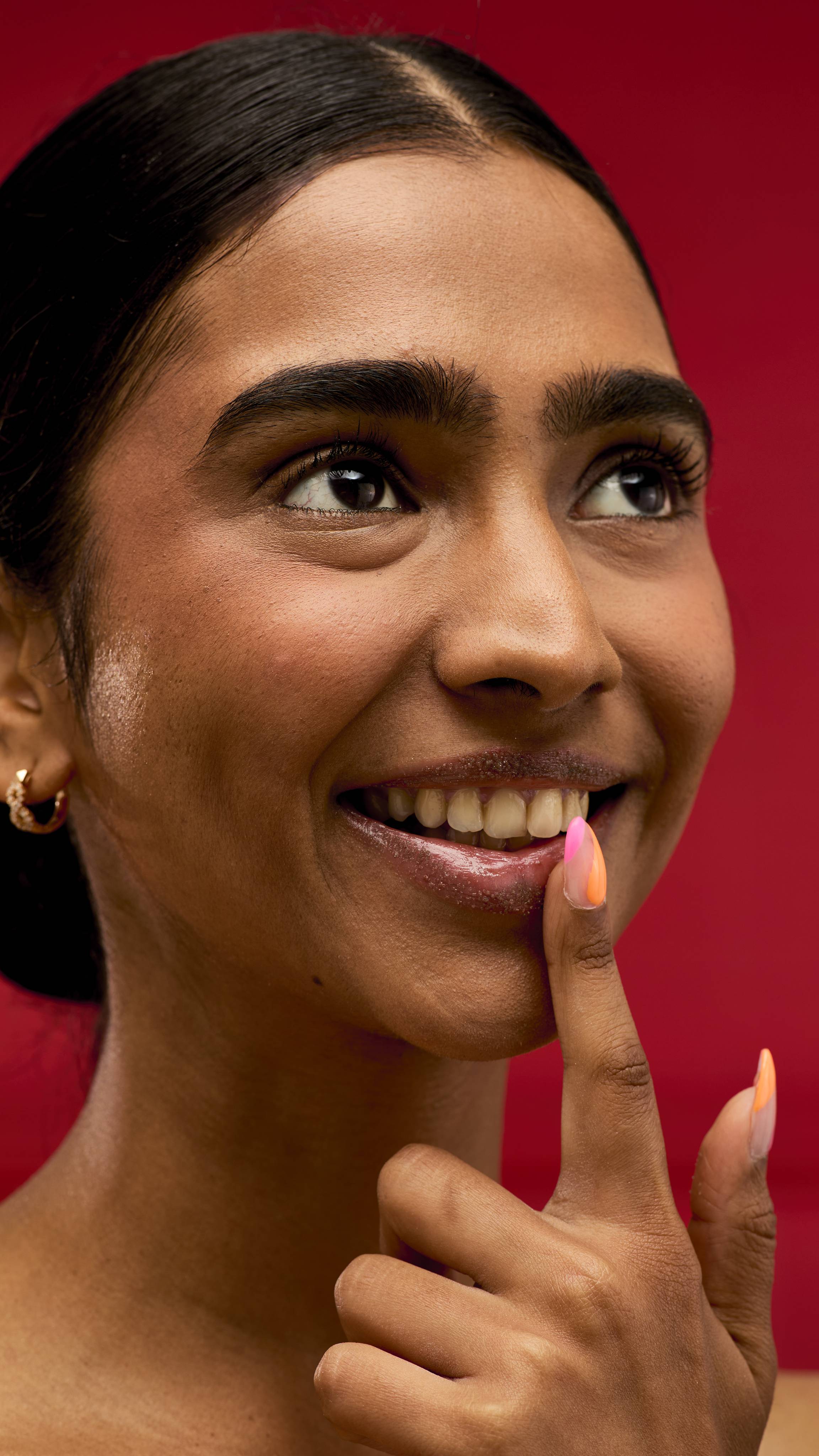 A smiling person looks away from the camera while gently touching their bottom lip with a finger. The person's lips glisten, suggesting the use of a LUSH After Dinner Mint lip scrub. The background is a vibrant red.