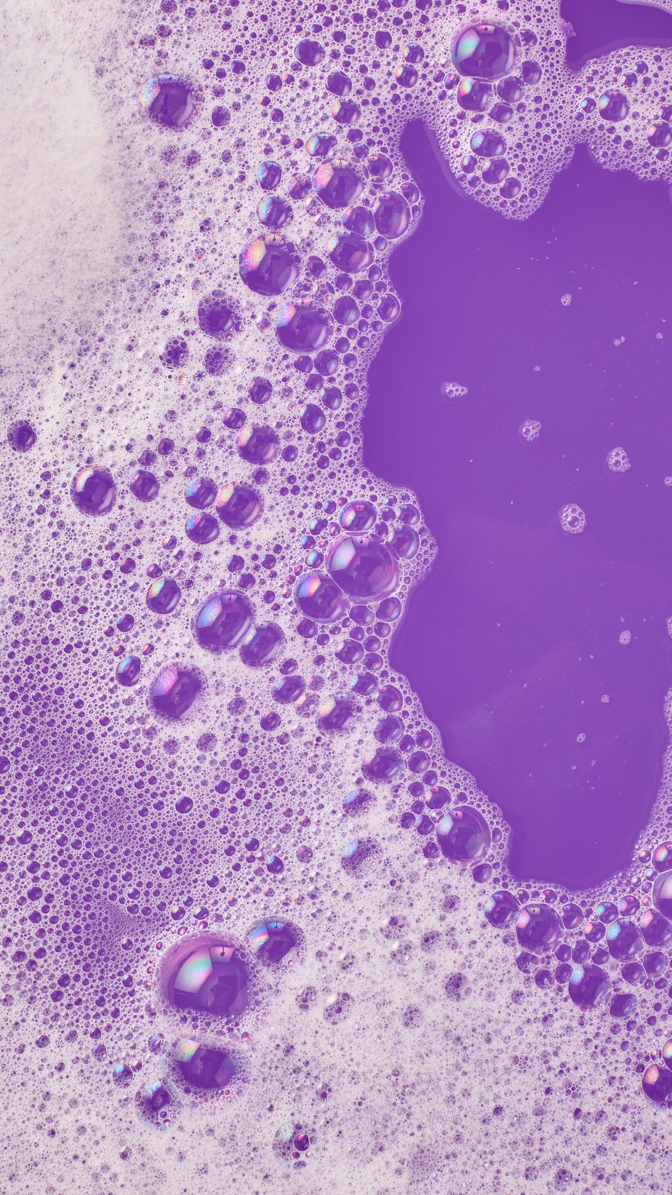 The image shows vibrant purple bath water largely covered in abundant white, frothy bubbles. Smooth, darker purple channels of water wind through the foam, creating an abstract, bubbly landscape from the 'Alien' bubble bar.