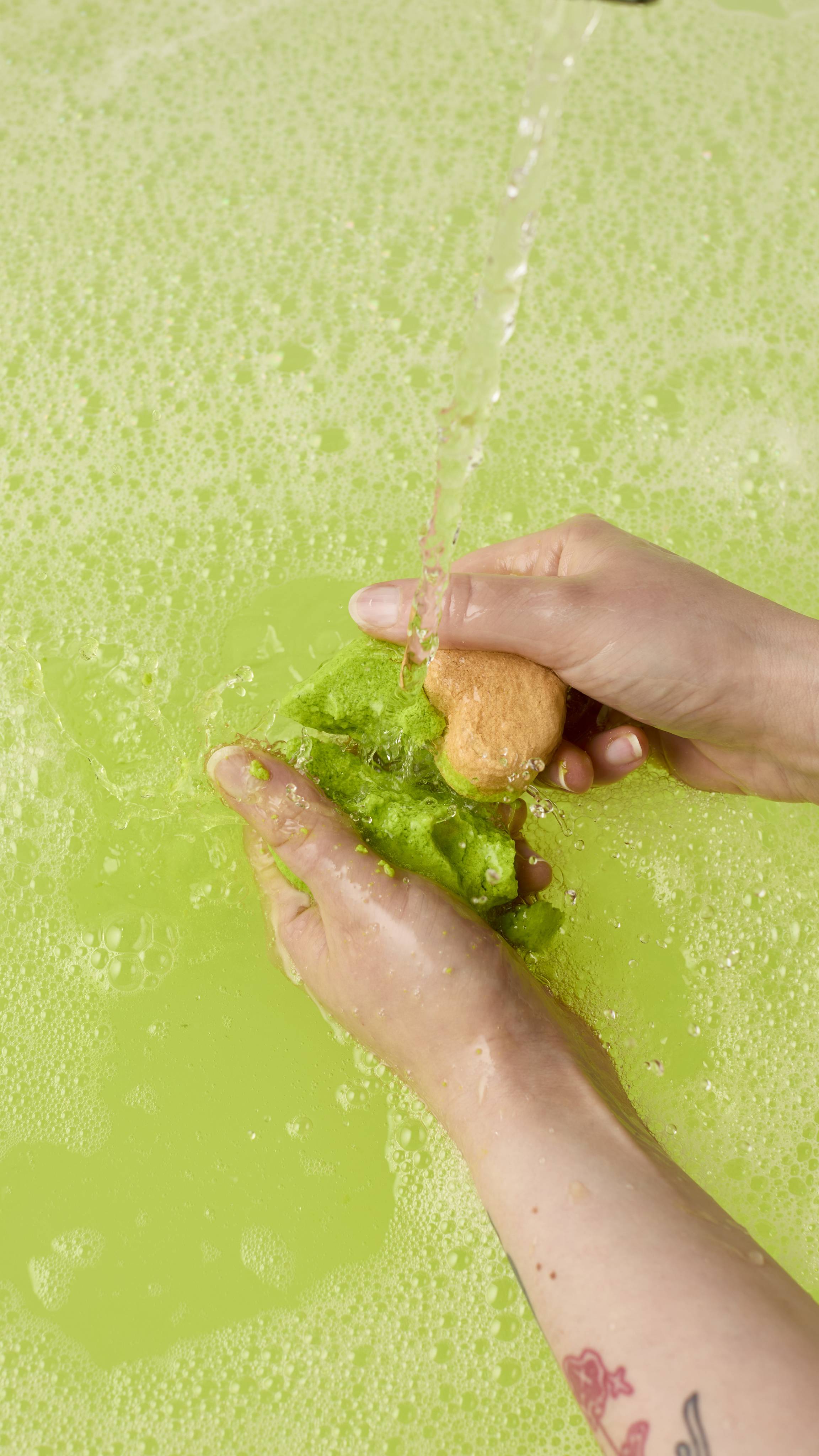 The image shows two hands crumbling a green "Avo Cuddle" bubble bar under a running tap in a bath. The product dissolves into the pale green water, creating a thick, foamy layer of white bubbles across the surface.