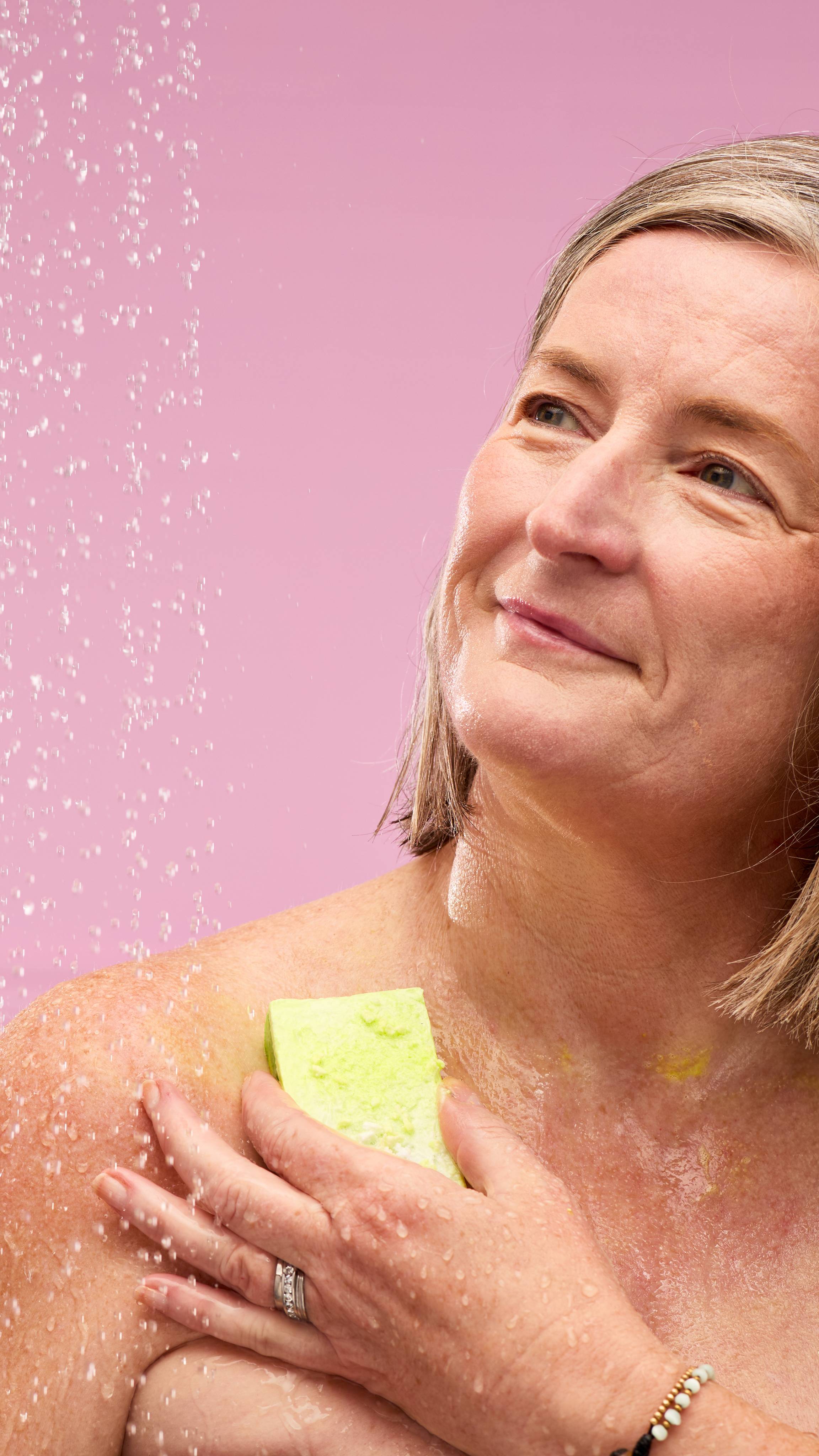 A fair-skinned person is shown from the chest up, in a shower against a pink background. They're smiling and looking up, with water droplets on their face and body. They are using the rectangular Baked Alaska soap slice to wash their shoulder.