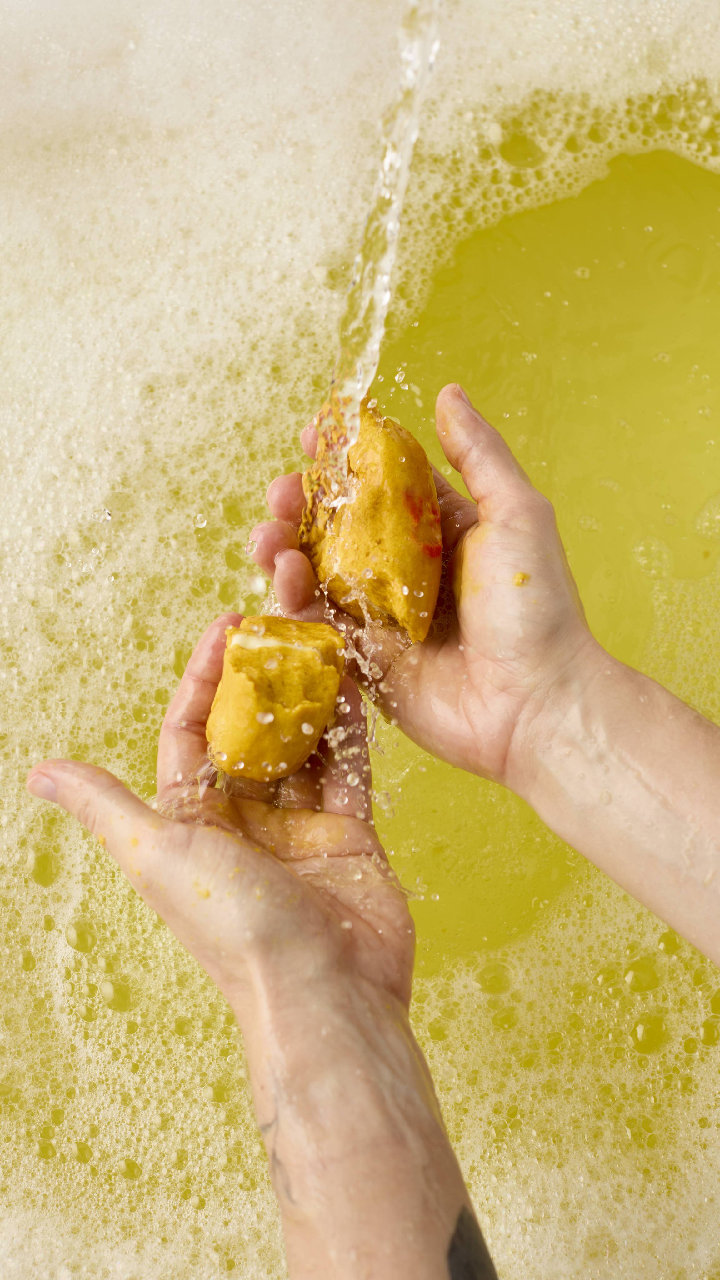 The image shows two hands crumbling the banana-shaped bubble bar under a stream of running water. The water splashes against the bar, creating pale yellow bubbles and foam in the bath below.