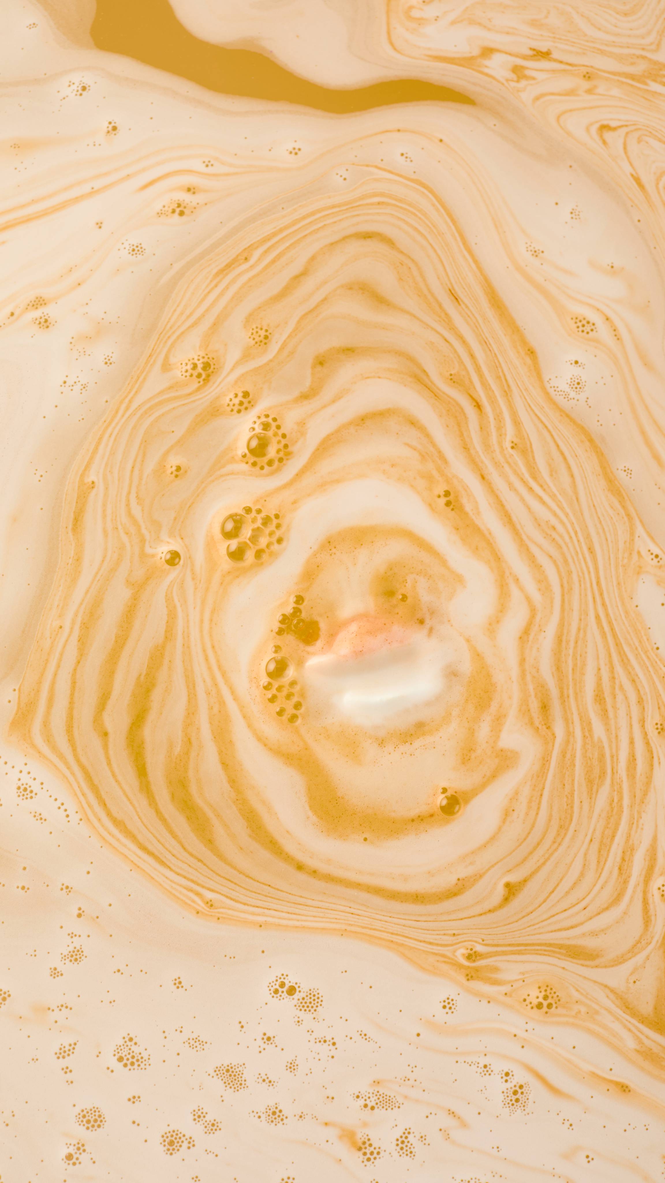 The Big Dilemma bath bomb dissolves in water, creating expanding ripples of thick, opaque orange and creamy white foam. The marbled pattern spreads across the surface, resembling a liquid silk texture.
