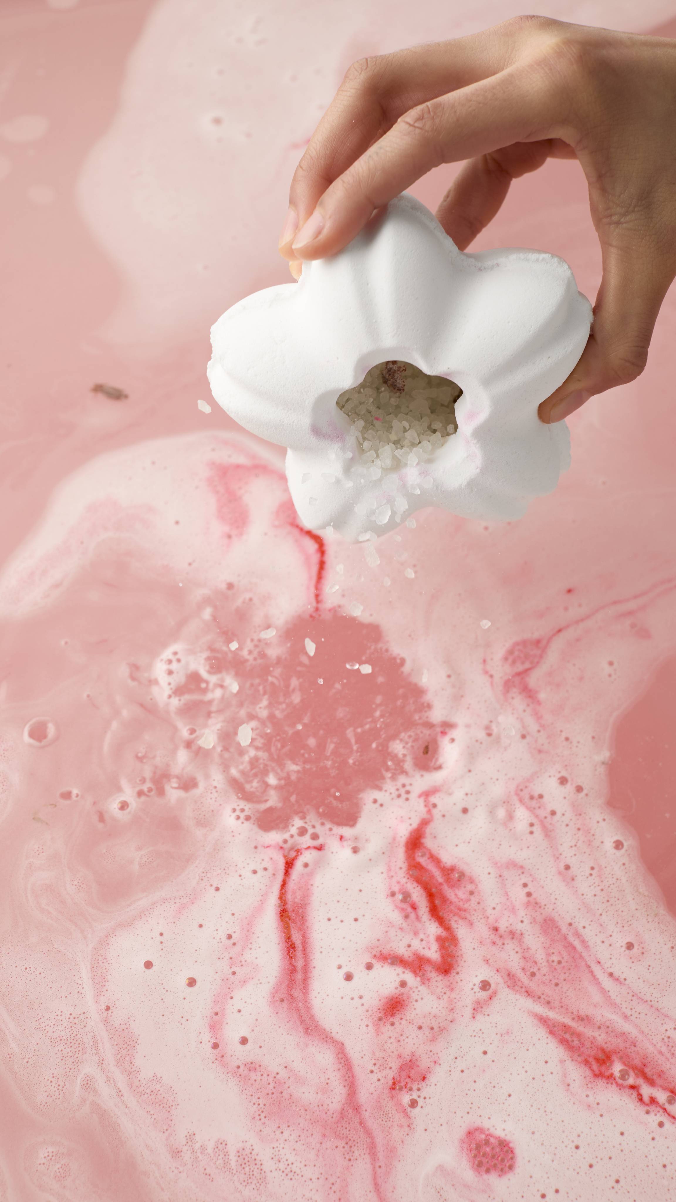 The image shows the model's hand tilting the Blooming Cherry Blossoms bath bomb, pouring out white bath salts into pink, swirling water. The small pink flower from the centre had been removed, revealing a hollow core filled with bath salts.