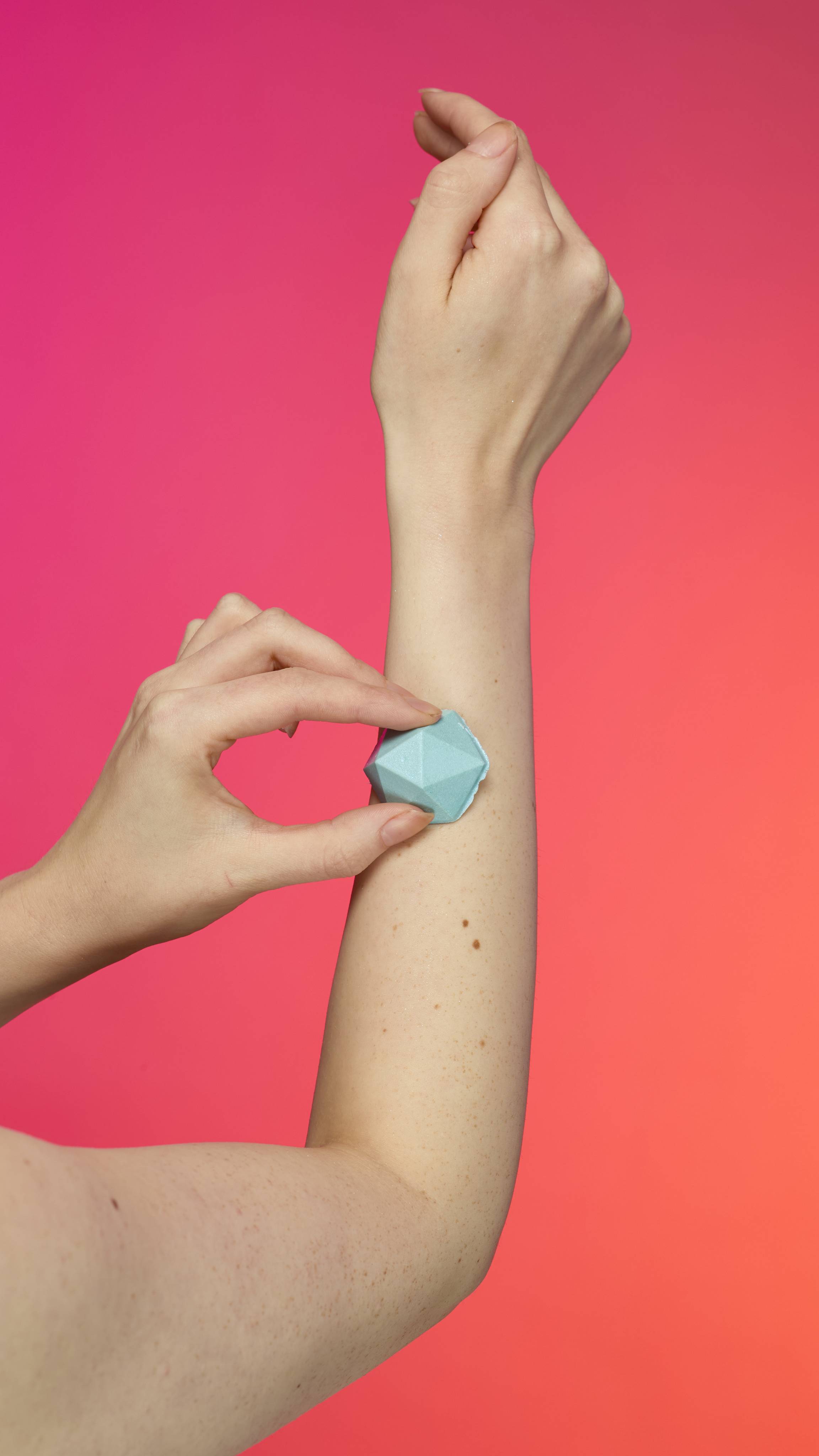 Two hands with fair skin apply the faceted, pyramid-shaped Blue glitterbug body bar to an arm. The skin has a subtle sheen of plastic free glitter against a vibrant gradient of magenta, pink, and orange.