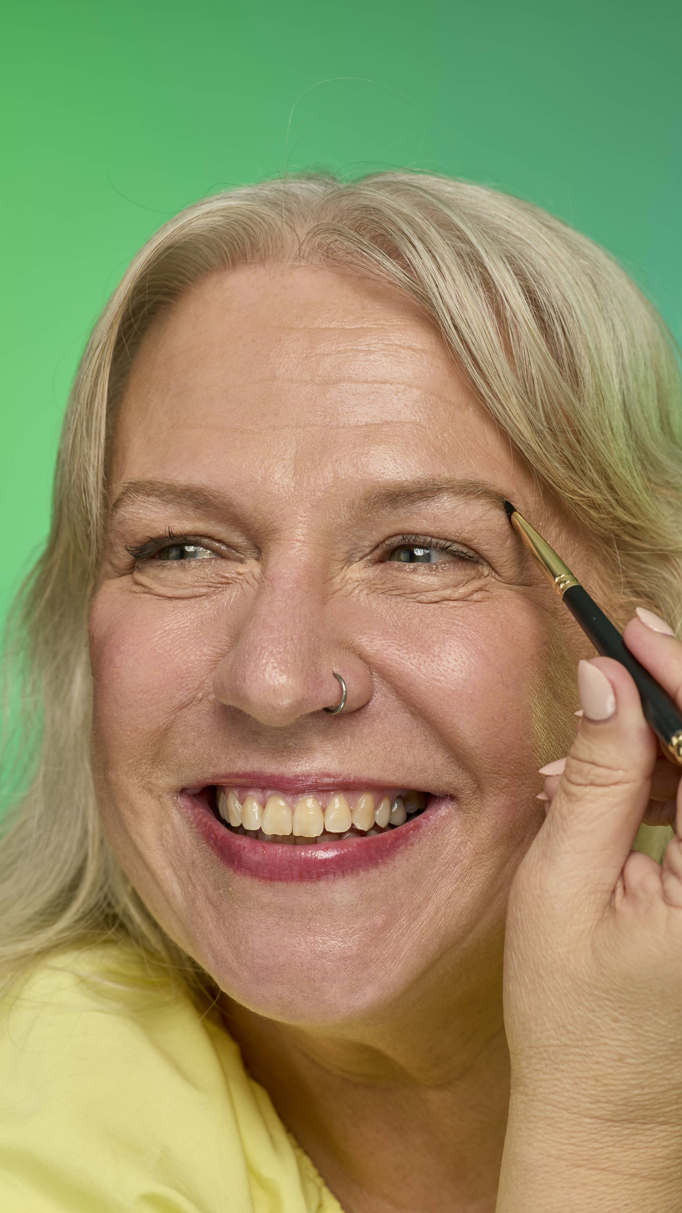 The image shows a model with blonde hair on a green background. They are smiling and holding an eyebrow brush up to their face. Their eyebrows look thick and full thanks to the Blonde brow pomade.
