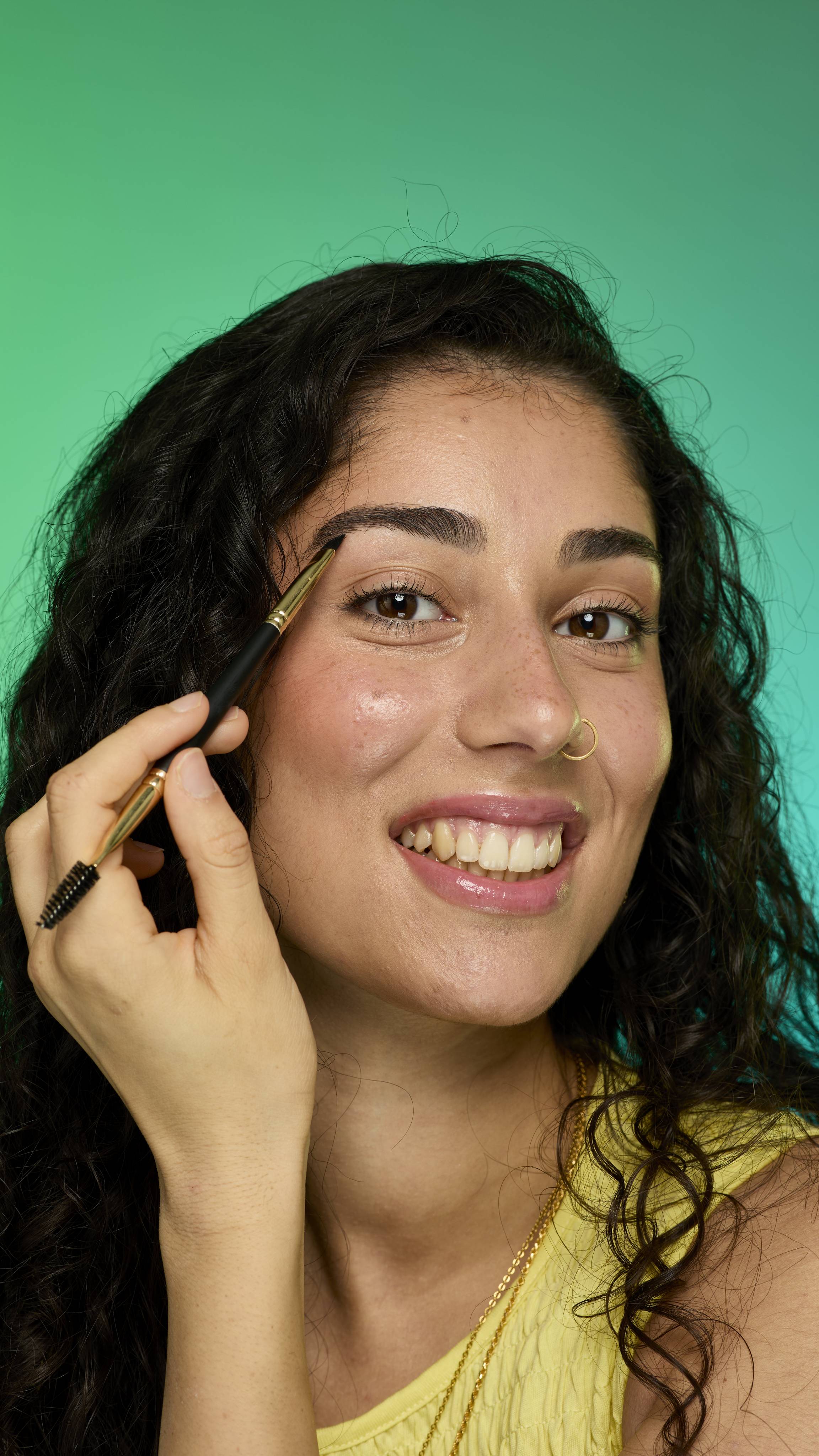 The image shows a model with dark curly hair on a green background. They are smiling and holding an eyebrow brush up to their face. Their eyebrows look thick and full thanks to the Brun brow pomade.
