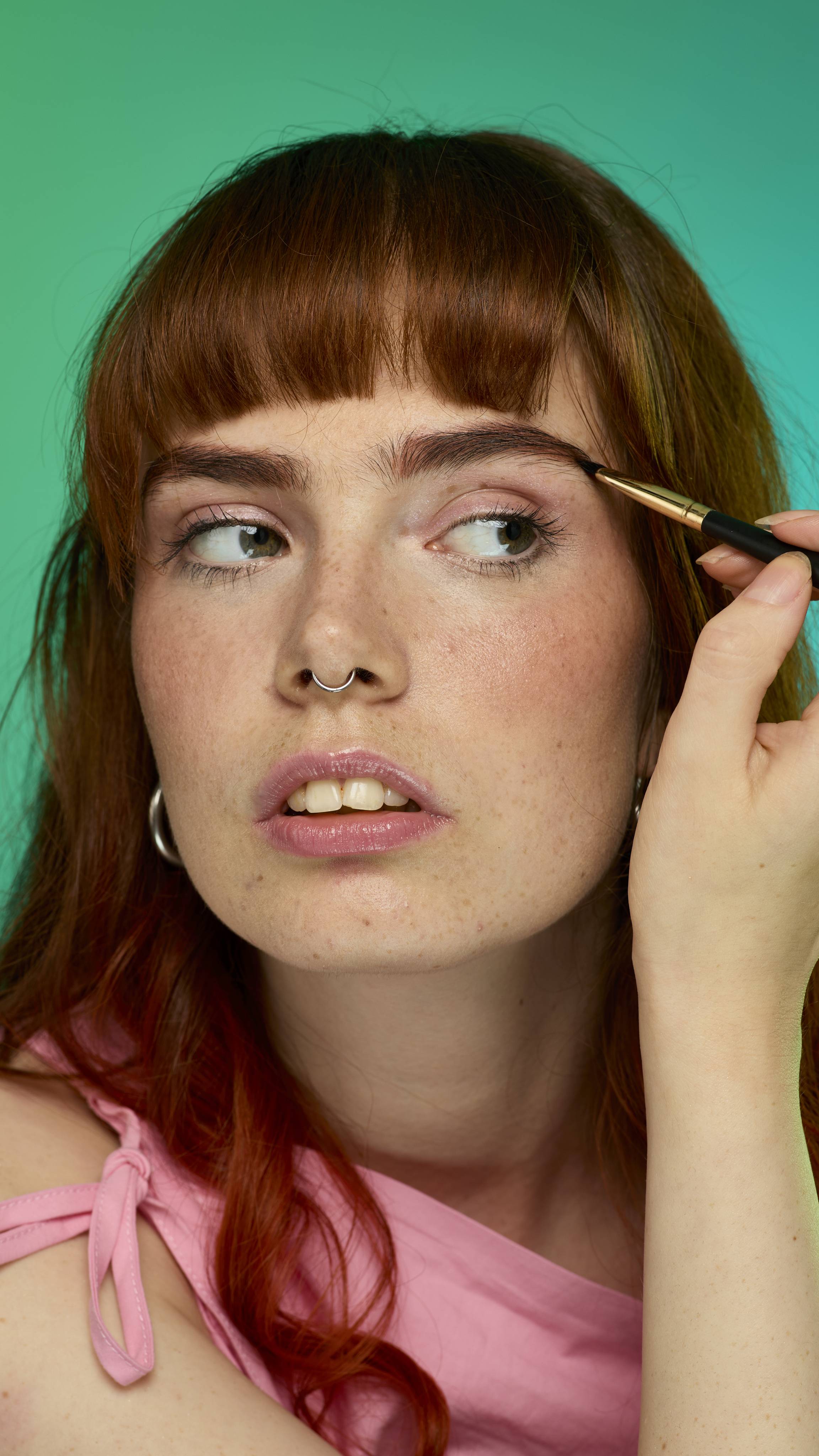 The image shows a model with fringed auburn hair on a green background. They are focusing and holding an eyebrow brush up to their face. Their eyebrows look thick and full thanks to the Rouge brow pomade.