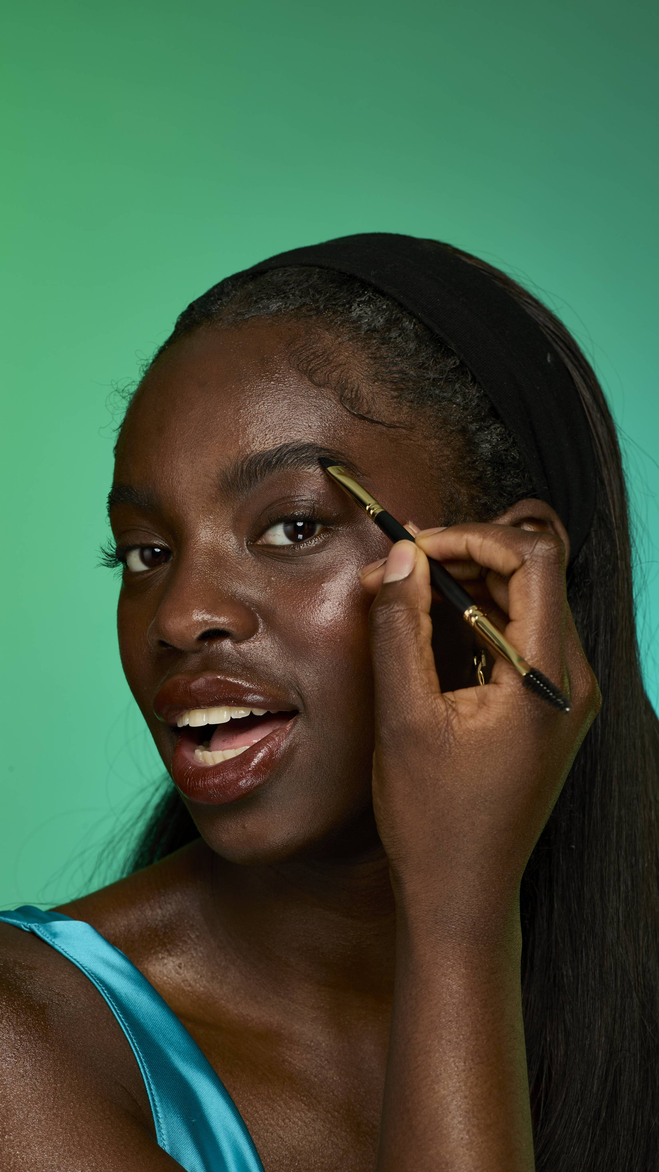 The image shows a model with dark straight hair on a green background. They are smiling and holding an eyebrow brush up to their face. Their eyebrows look thick and full thanks to the Smoke brow pomade.