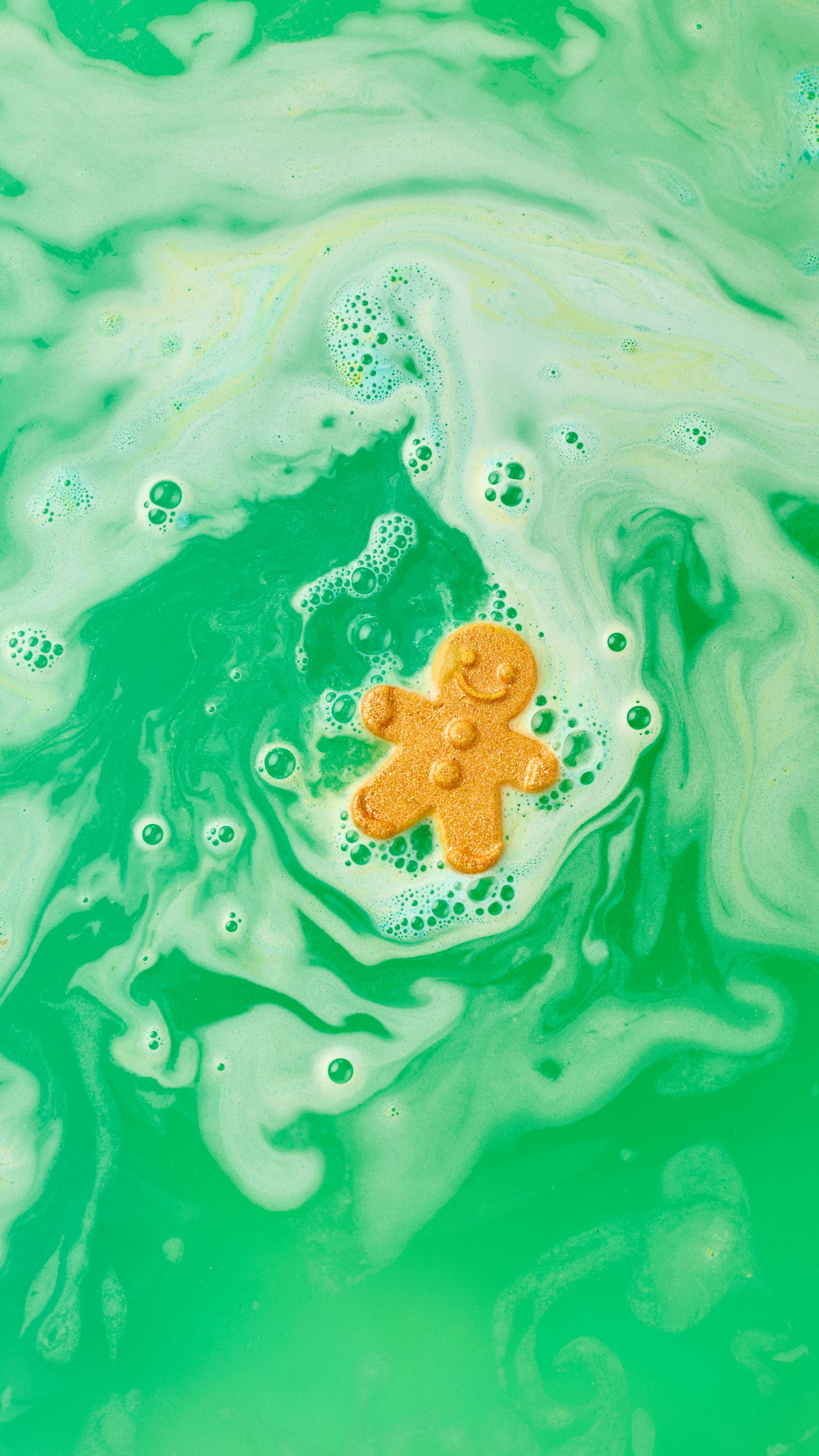 The image shows a golden, gingerbread man-shaped, Catch Me If You Can bath bomb fizzing in swirling green and yellow bath water. The water forms a colourful spiral around the dissolving product, blending with foam and bubbles.