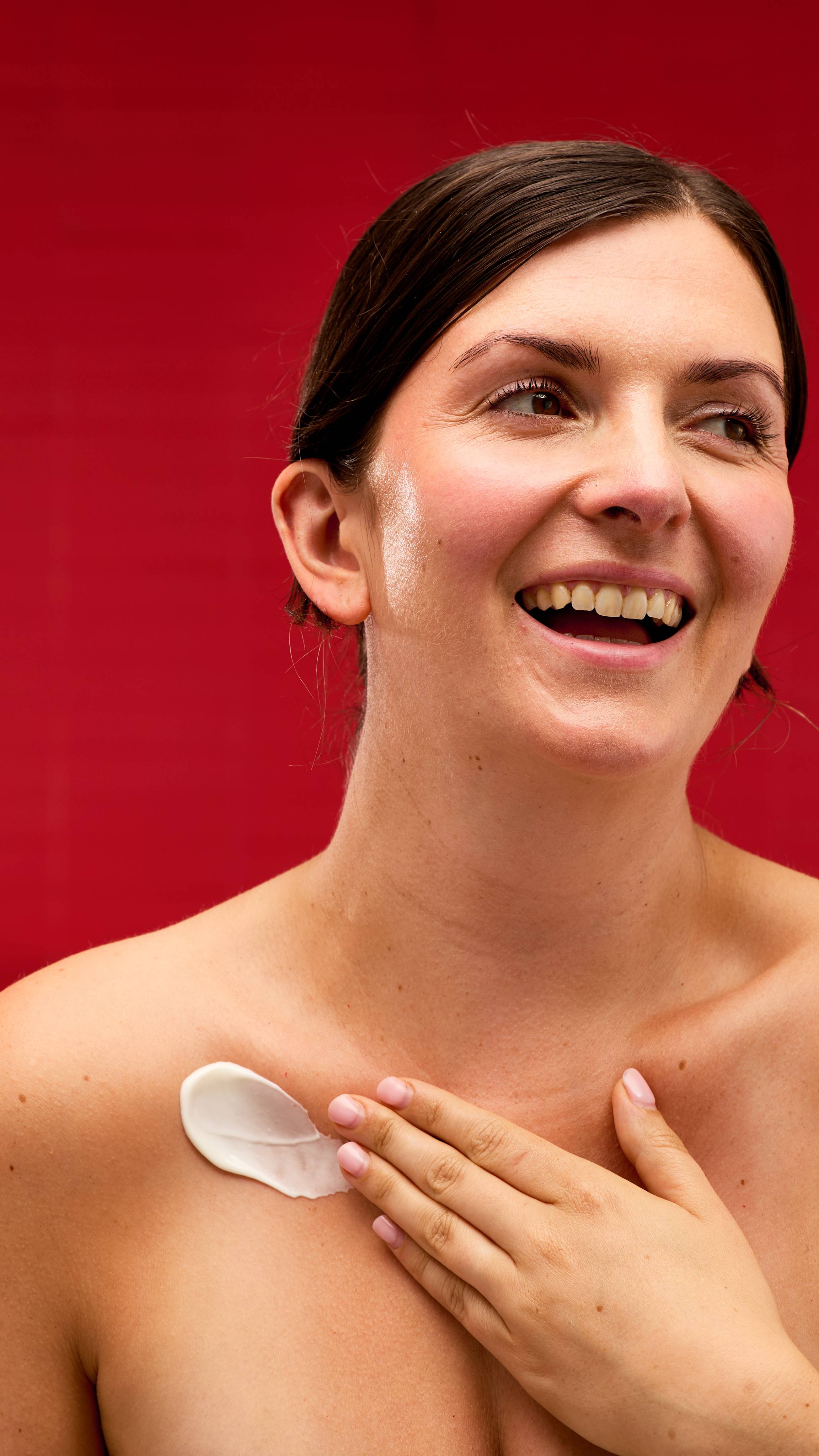 The image shows a solid red background. A model can be seen from the chest up as they are smiling while applying the thick, creamy Celebrate body lotion to their collarbone. 