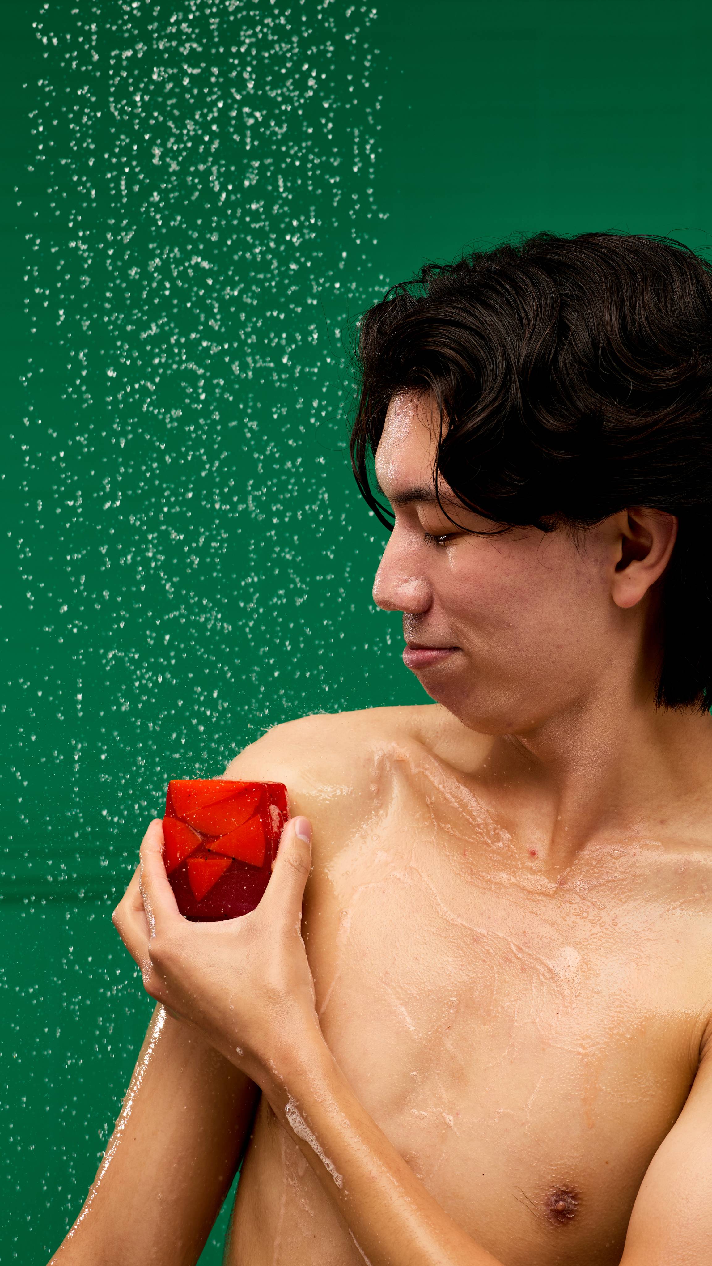 A person is shown from the chest up, in a shower against a green background. They're smiling and looking up, with water droplets on their face and body. They are using the red, Christmas Cranberry soap slice to wash their shoulder.