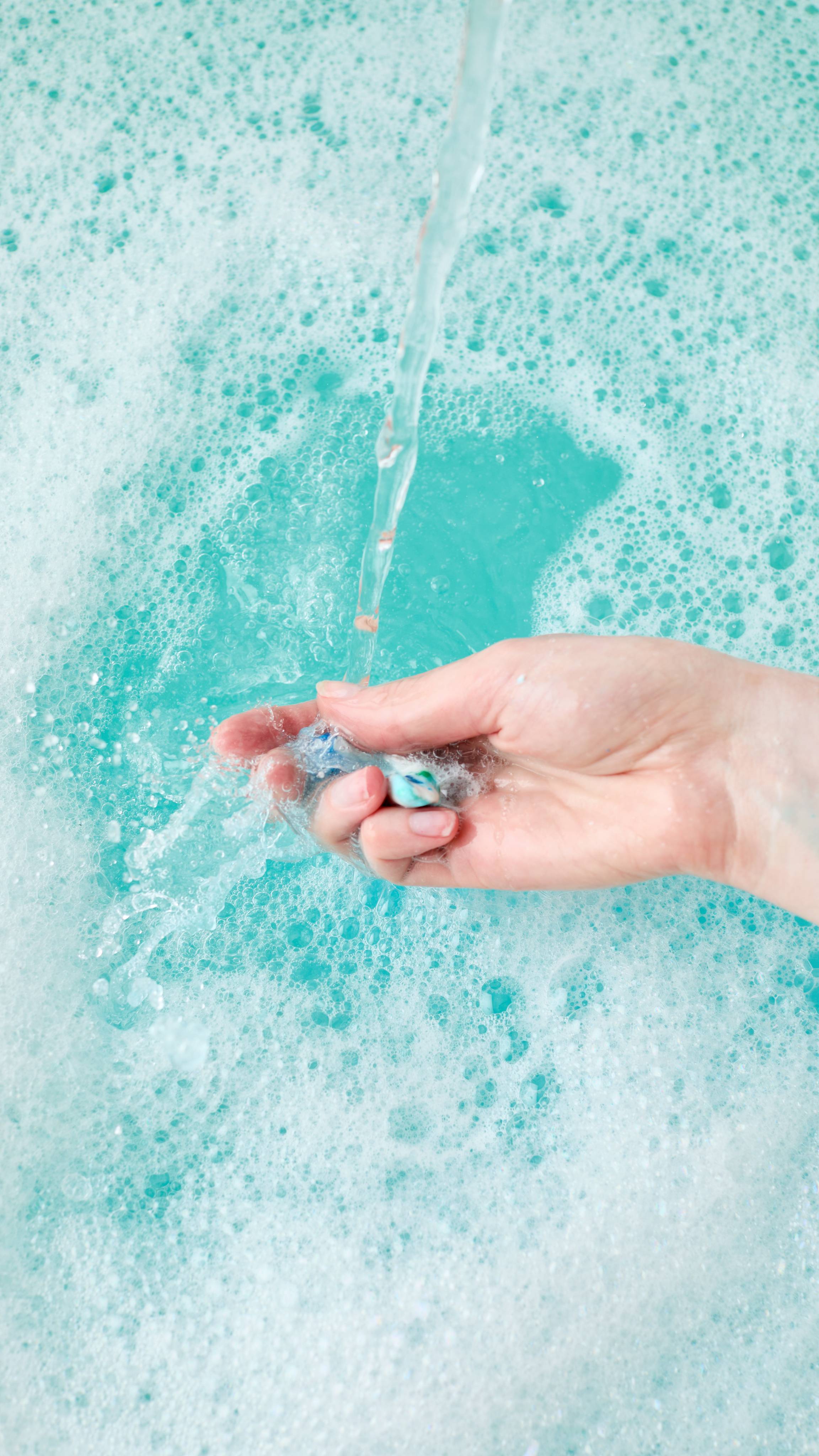 A fair-skinned hand gently squeezes and crumbles some of the Christmas Lights Fun product under a stream of water, creating teal-coloured water and white foam in a bathtub.