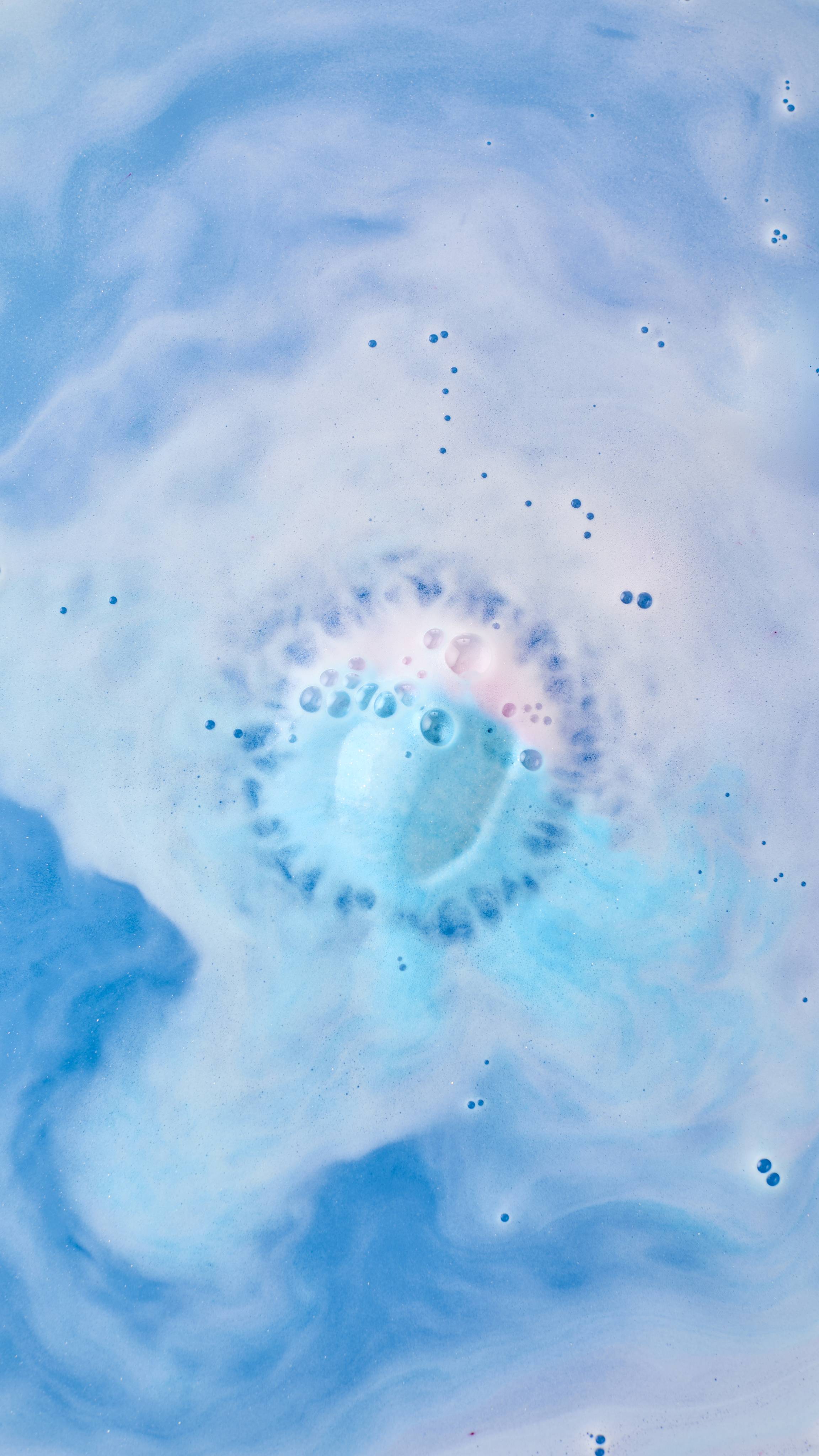 An aerial view of a bathtub filled with blue-tinted water and swirling white foam. In the centre, a partially dissolved "Dad Dancing" bath bomb creates a concentrated circle of blue fizz and tiny bubbles as it releases colour into the bath.