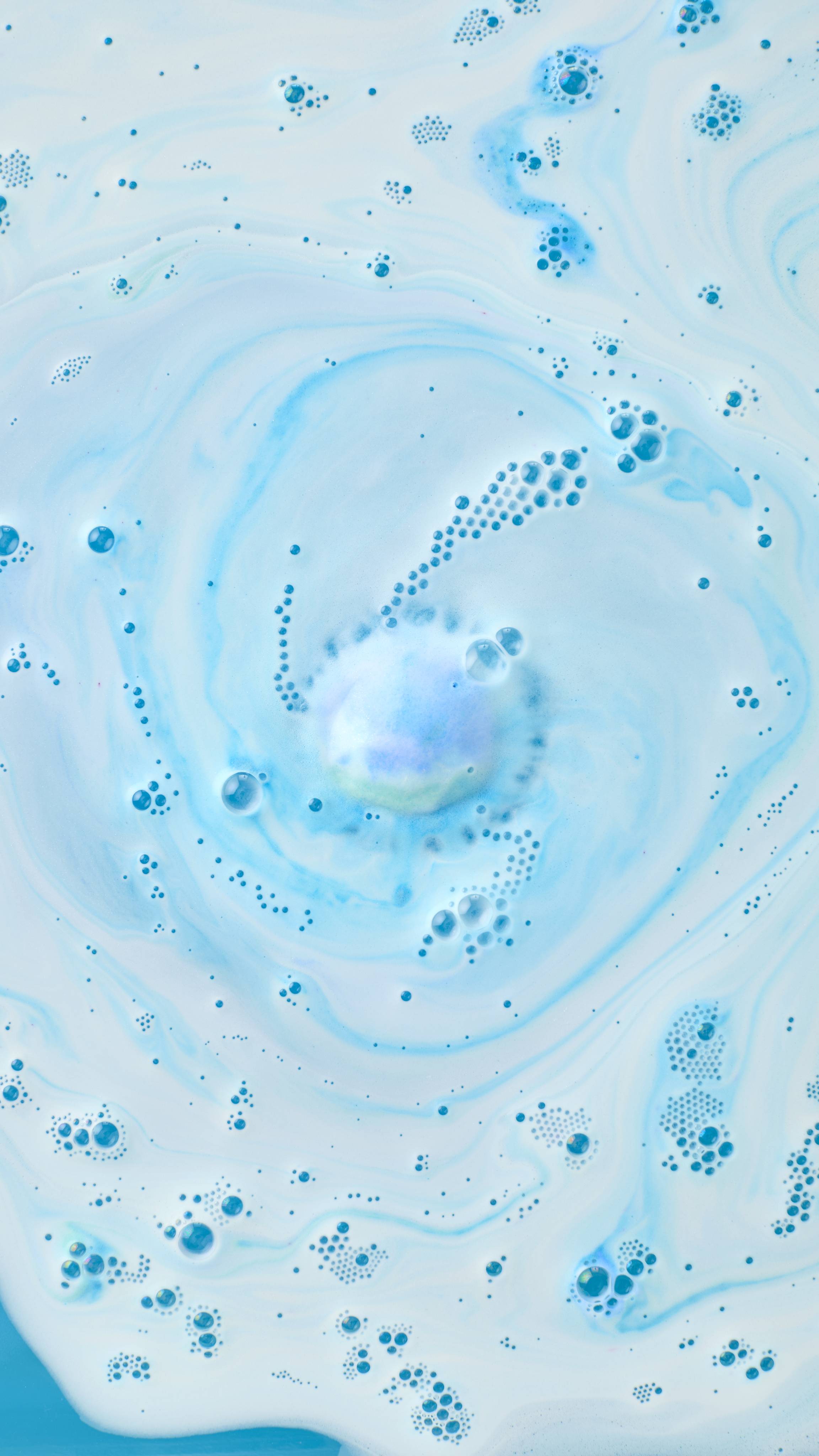 An aerial view of blue and white bath foam from the "Easy Breezy" bath bomb, swirling into a soft, hypnotic spiral. In the centre, the product dissolves, releasing delicate bubbles and a pale green core into the surrounding pale blue water.