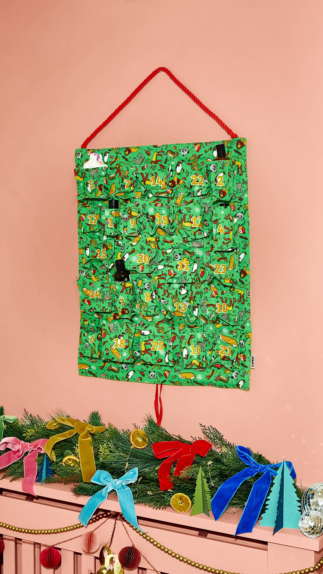 A green fabric Lush Advent calendar hanging by a red cord against a pink wall, with small products peeking out from numbered pockets, positioned above a mantelpiece decorated with festive greenery and colourful bows.