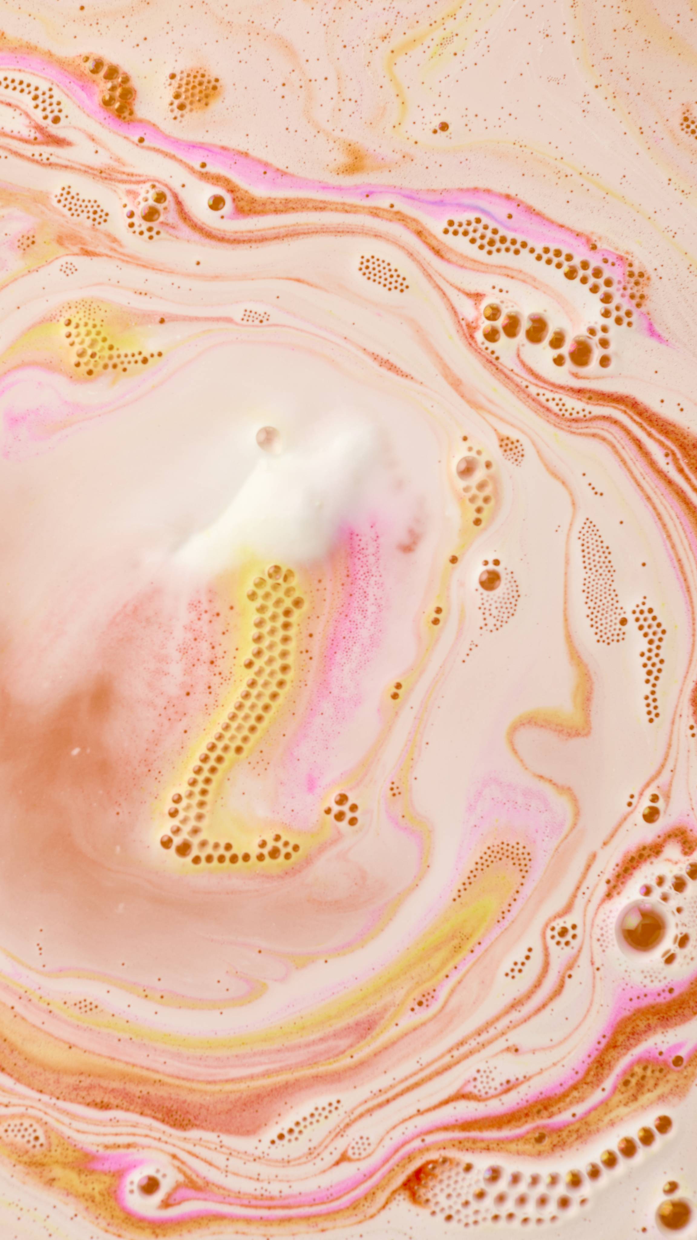 A close-up of the Follow the White Rabbit bath bomb as it dissolves in bathwater. Swirls of orange, pink, and yellow foam expand across the surface of the water, creating a vibrant, marbled sunset effect.