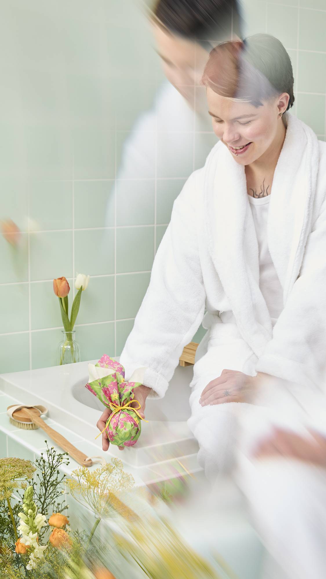 The image shows a model with short hair, in a white robe, sitting on the edge of a tiled bath. They are holding a bath bomb wrapped in the "Garden in Spring" Lokta paper gathered on top and tied with yellow twine.