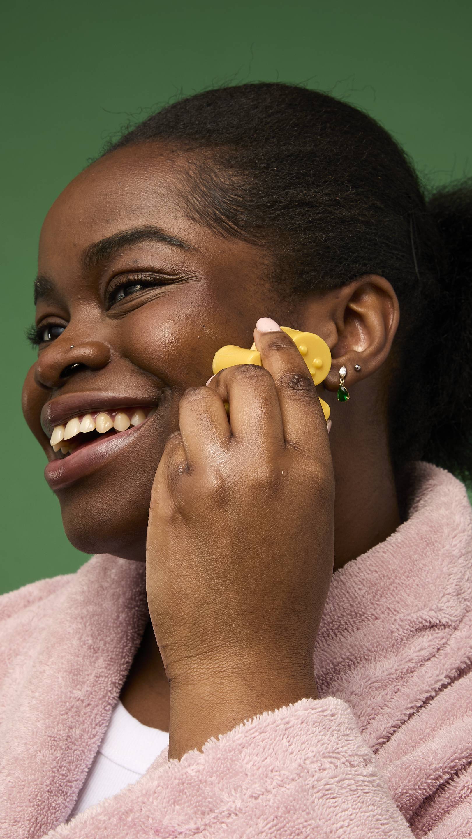 The image shows a model with dark skin, wearing a pink bathrobe from the chest up against a solid green background. They are rubbing a golden-yellow Gingerbread gummy facial wash over their cheek.