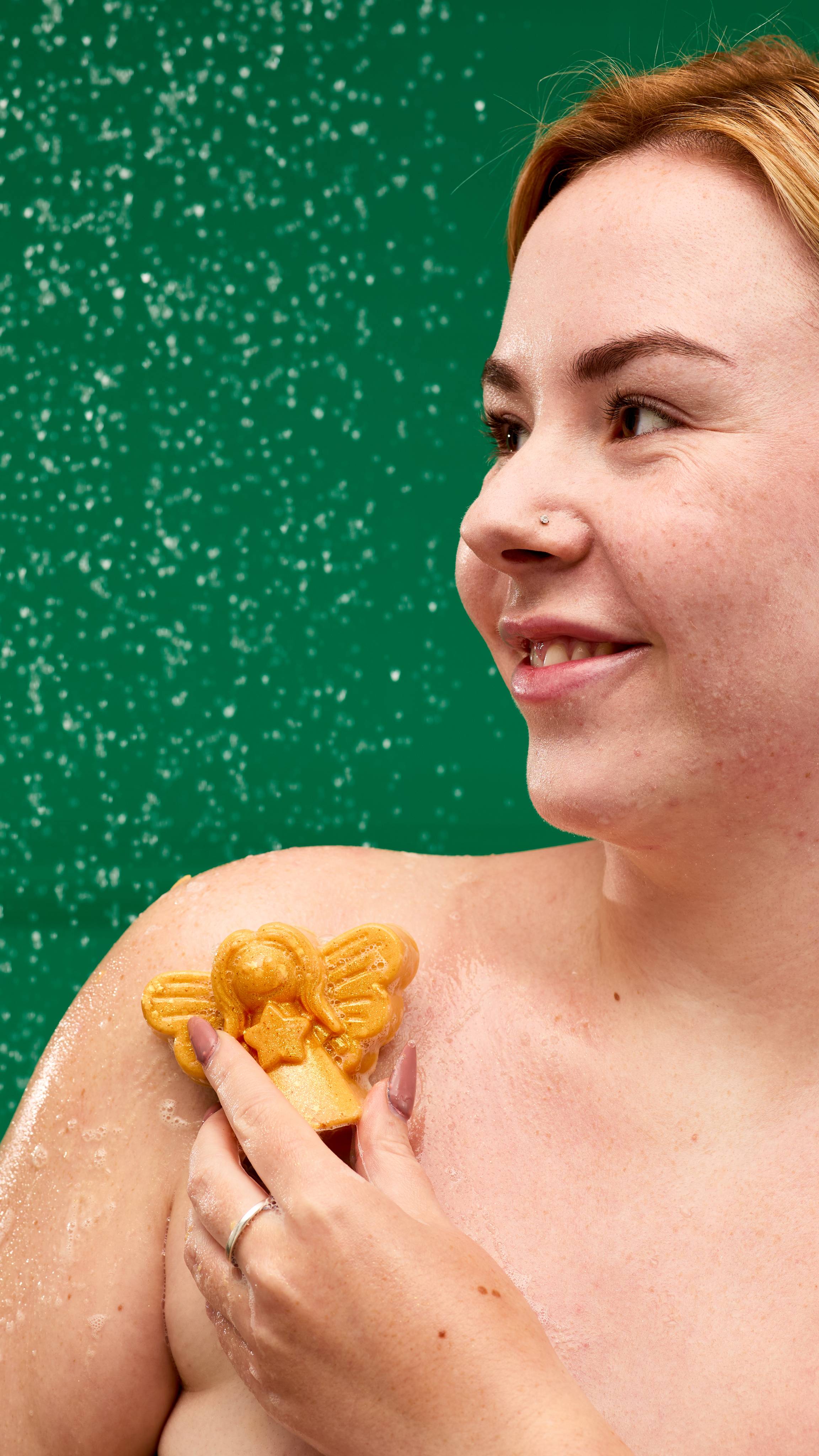 A person is shown from the chest up, in a shower against a green background. They're smiling and looking up, with water droplets on their face and body. They are using the white Golden Angel Delight soap to wash their shoulder.