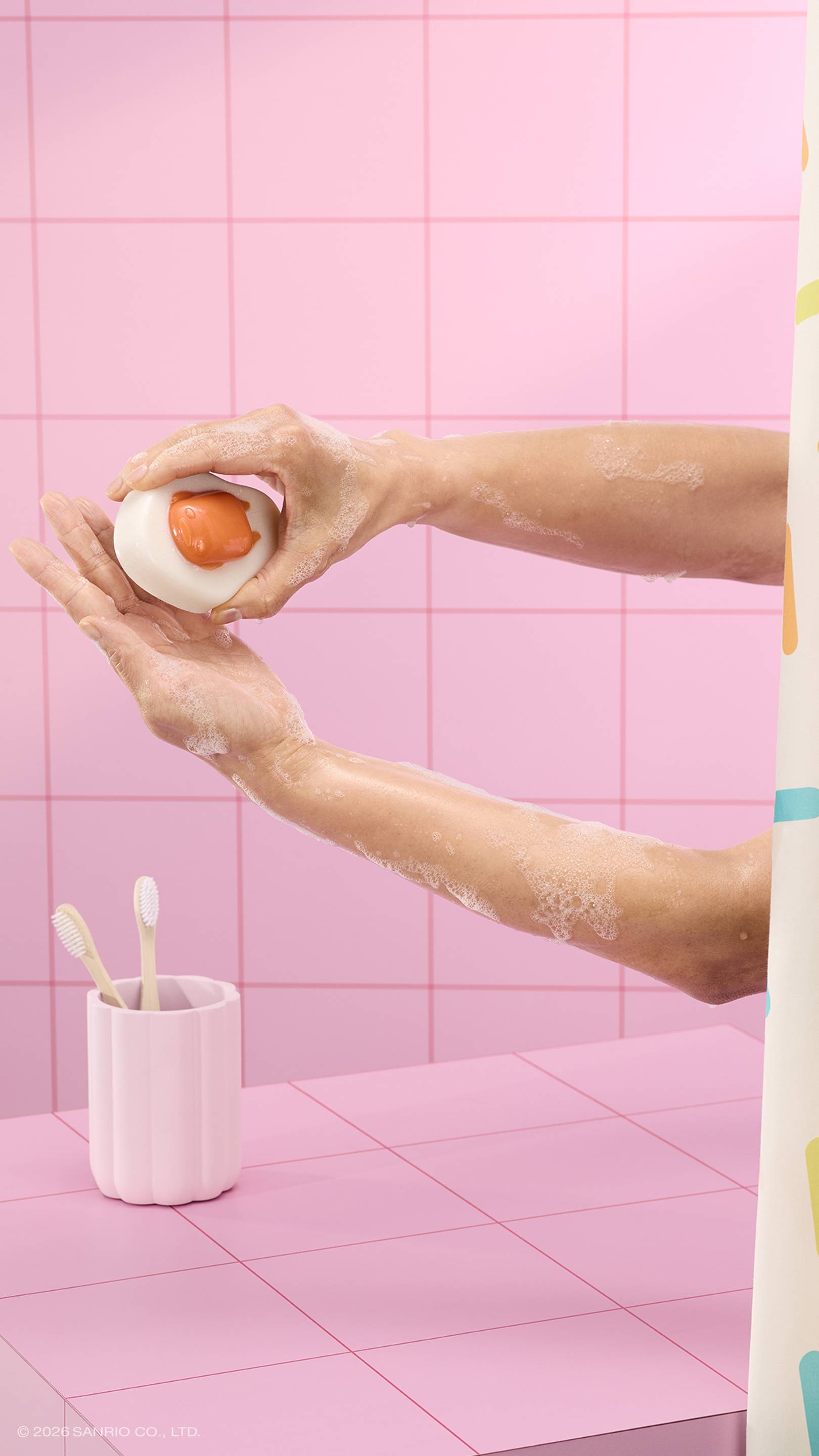 A person lathers their soapy arms using a glossy, egg-shaped "Gudetama" shower jelly in a pink tiled bathroom. A pink toothbrush holder and a sprinkle-patterned curtain are visible.