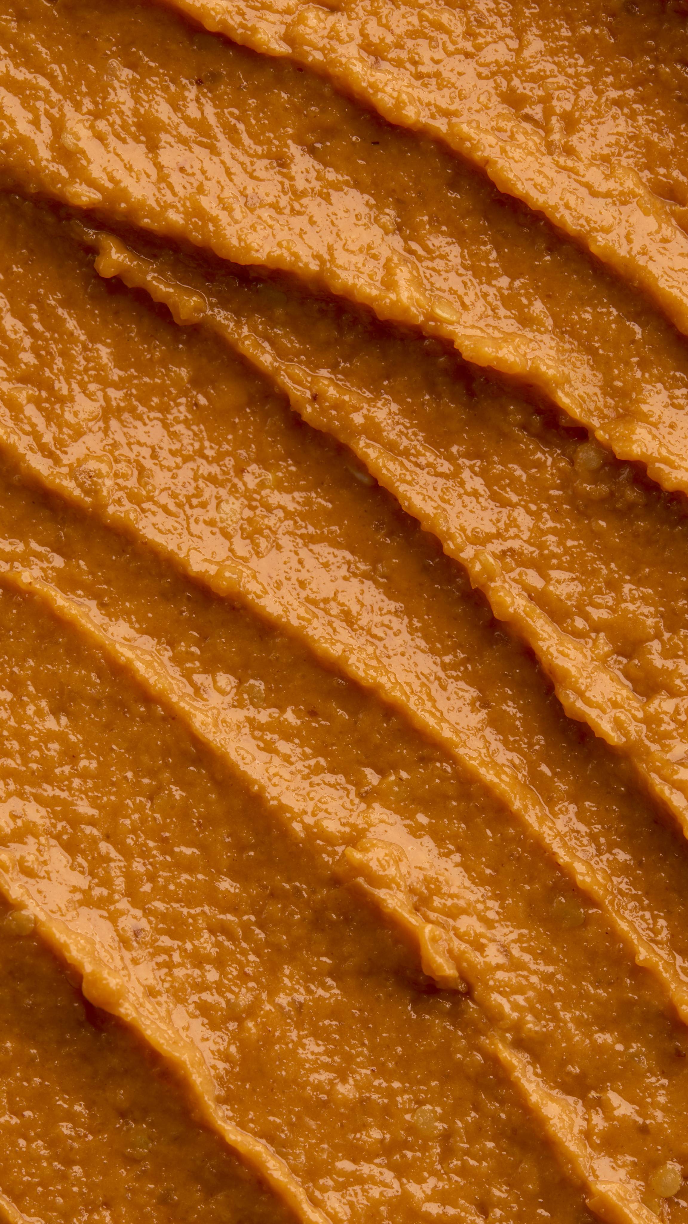 A textured, close-up macro shot of thick diagonal smears of bright orange Halloween Queen face mask. The rough paste features distinct ridges from application, filled with numerous tiny exfoliating specks.