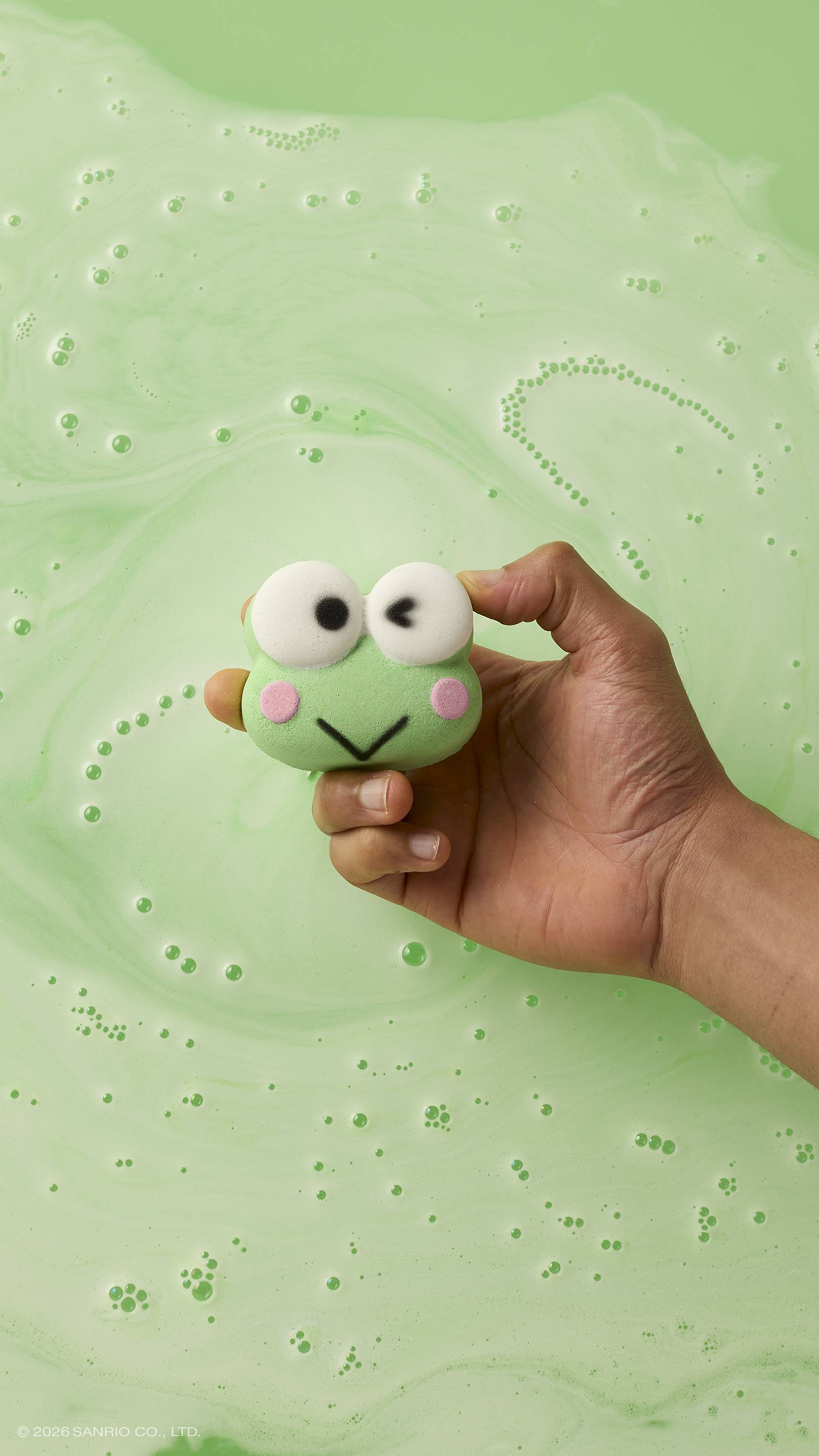 A hand holds a green, winking frog-shaped bath bomb over a bath of lime-green water. The bath bomb has pink cheeks and large white eyes, and is surrounded by swirling white foam and small bubbles on the water's surface. 