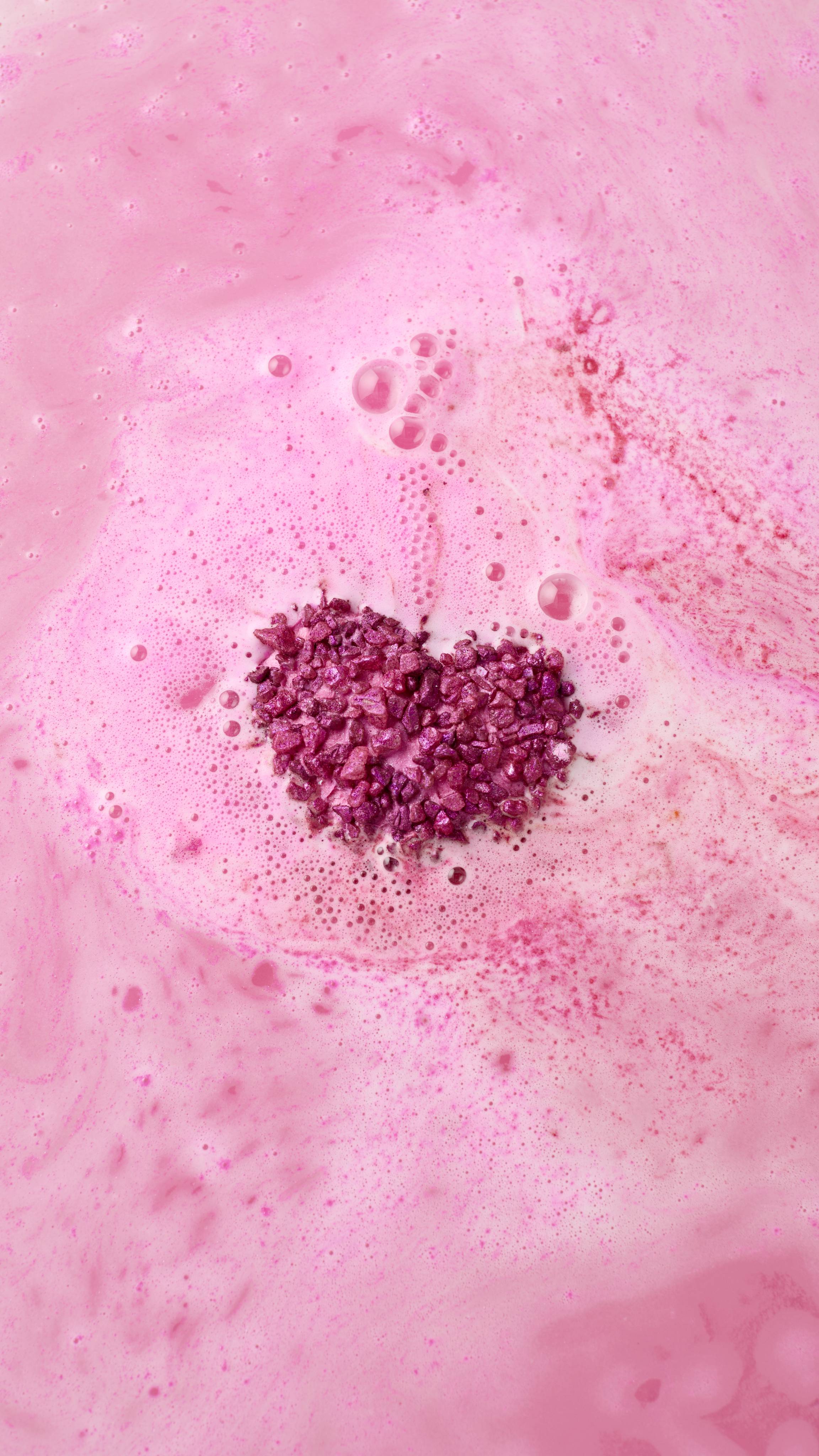 The heart-shaped Life In Rosy Light bath bomb has almost entirely dissolved into the bath. A cluster of sparkling magenta salt crystals remains in the centre, surrounded by a frothy layer of bubbles and vibrant pink water.