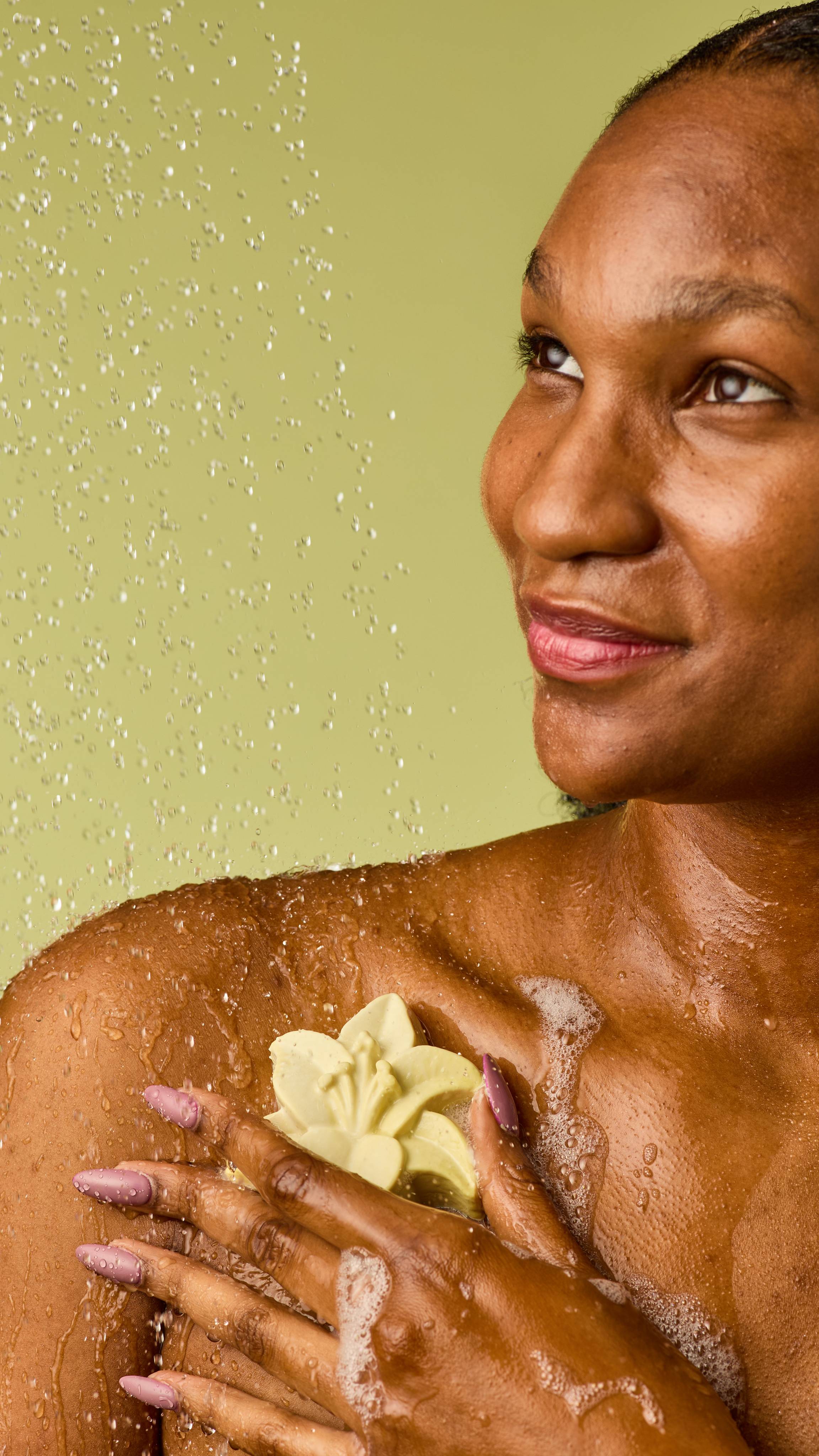 The image shows a person with a joyful expression, holding a pale cream, lily-shaped soap on their chest. Their hands, with long, light purple nails, are covered in water droplets and bubbles, against a light green background.