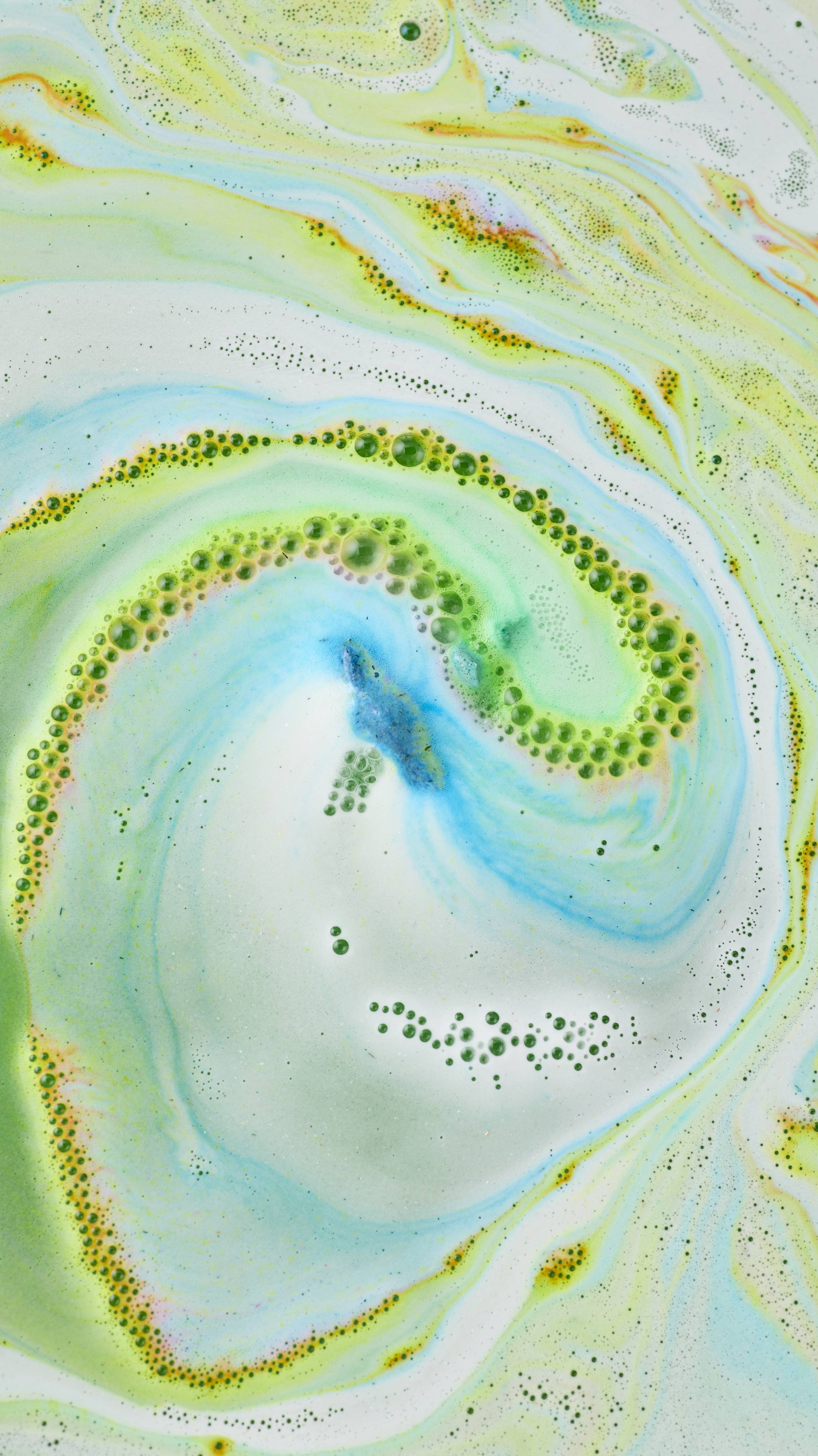 An aerial, abstract view of swirling bath art created by the "Lola's Aurora Borealis" bath bomb. Vibrant lime-green and turquoise-blue foam spirals together with white suds, creating a marbled, whirlpool effect. 