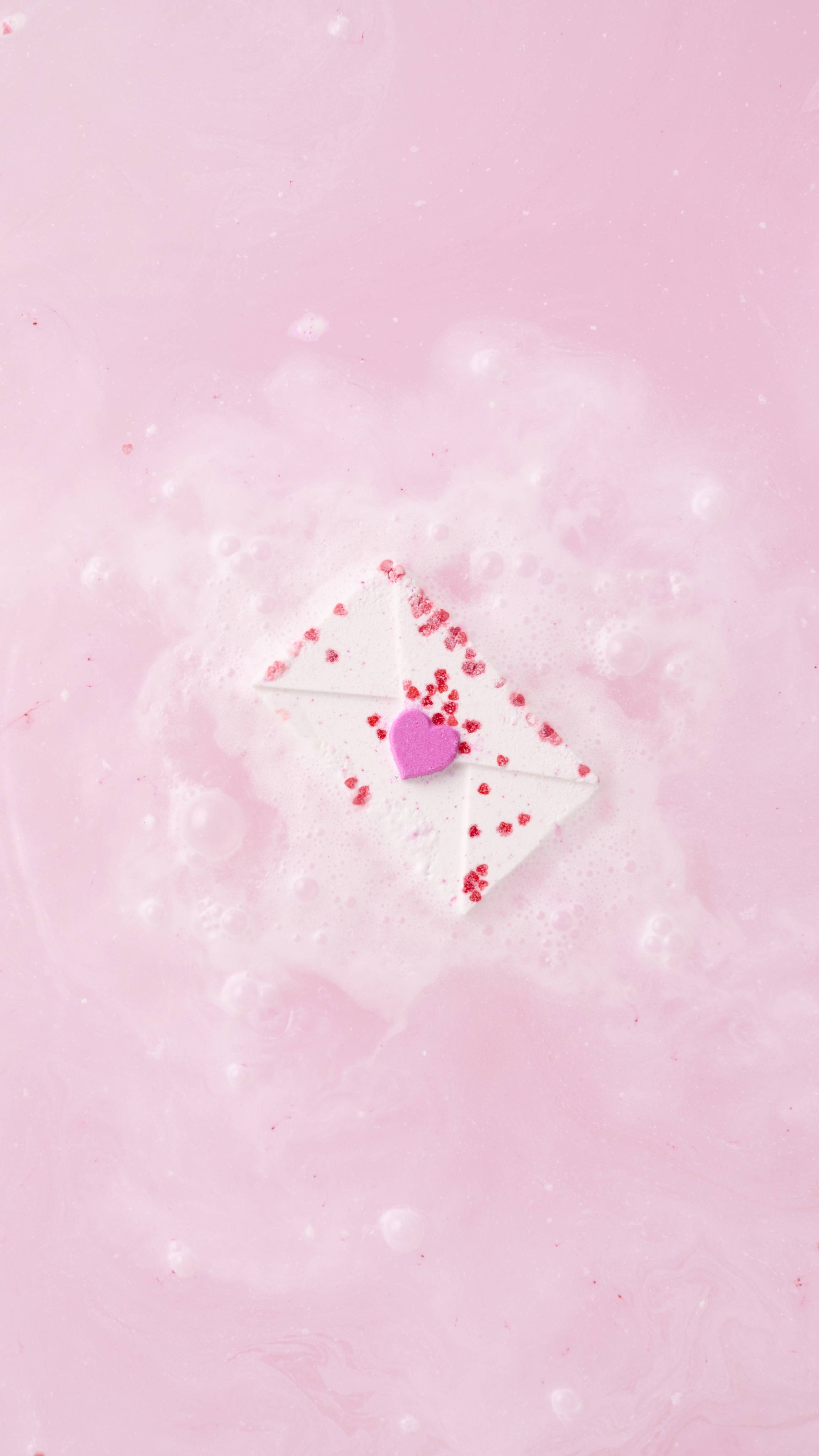 The envelope-shaped Love Letter bath bomb floats in the centre of a bath, surrounded by a cloud of white and pale pink fizz. The water around it is a milky, pastel pink with delicate bubbles on the surface.