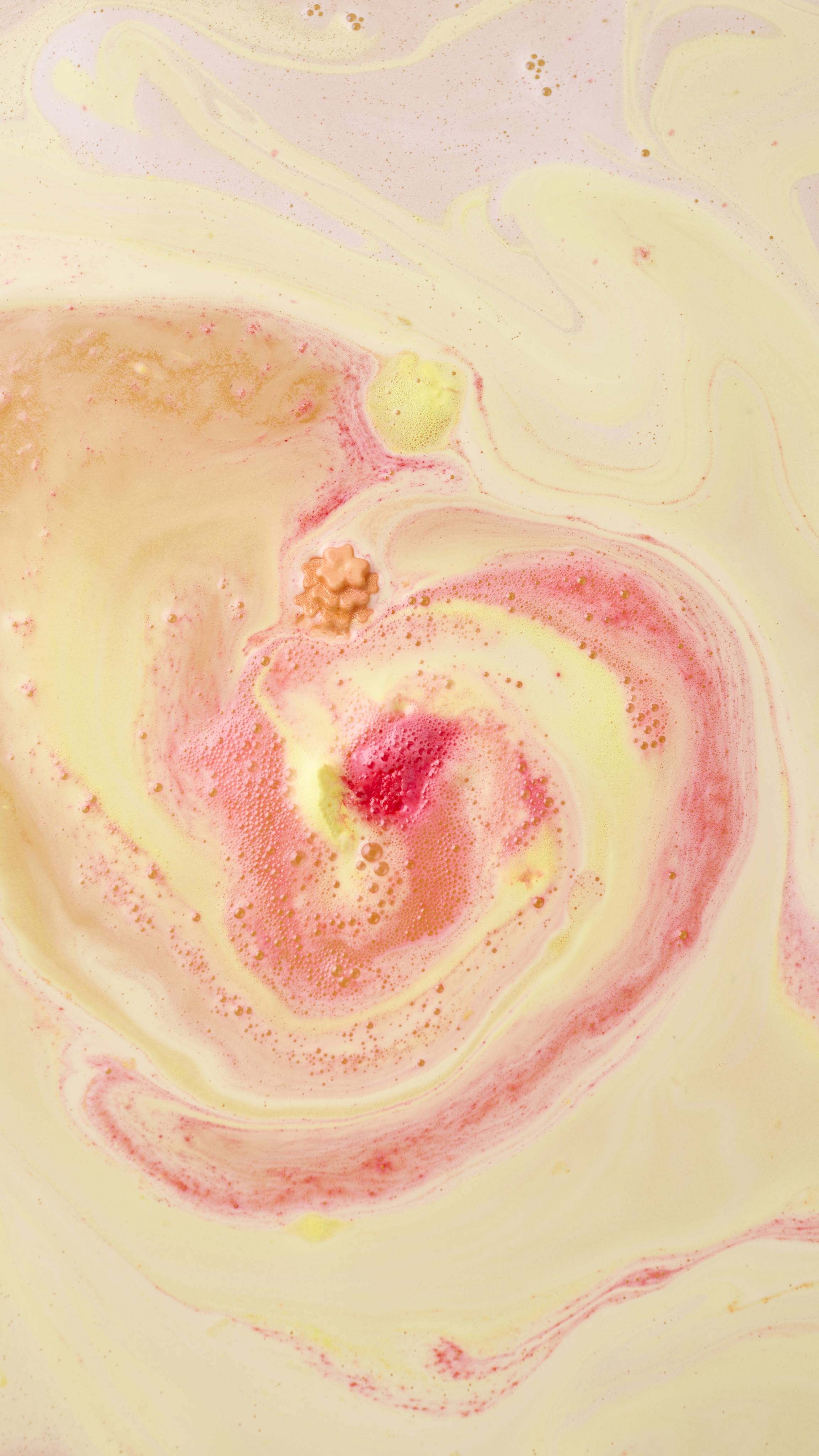 An aerial view of a yellow and pink "Marigold Tea" bath bomb dissolving in water, creating a vibrant, swirling pattern of creamy pastel yellow and bright pink foam. The colours marble across the surface of the bathwater.