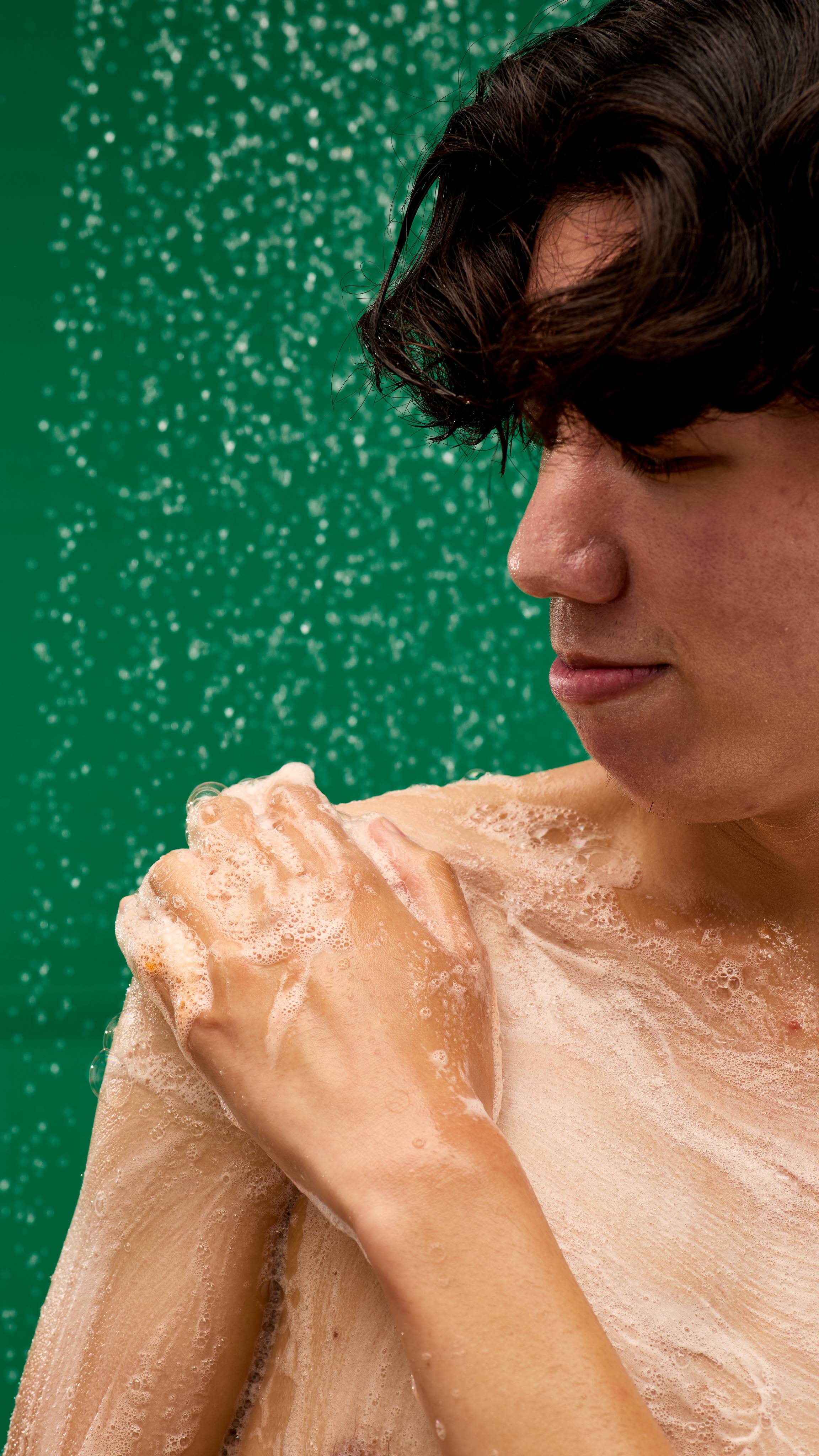 A person with their eyes closed in contentment lathers up their shoulder with a white foam created by LUSH Merry Christmas Everyone shower gel. Water droplets are visible in the air, creating a sparkling effect against a vibrant green background.