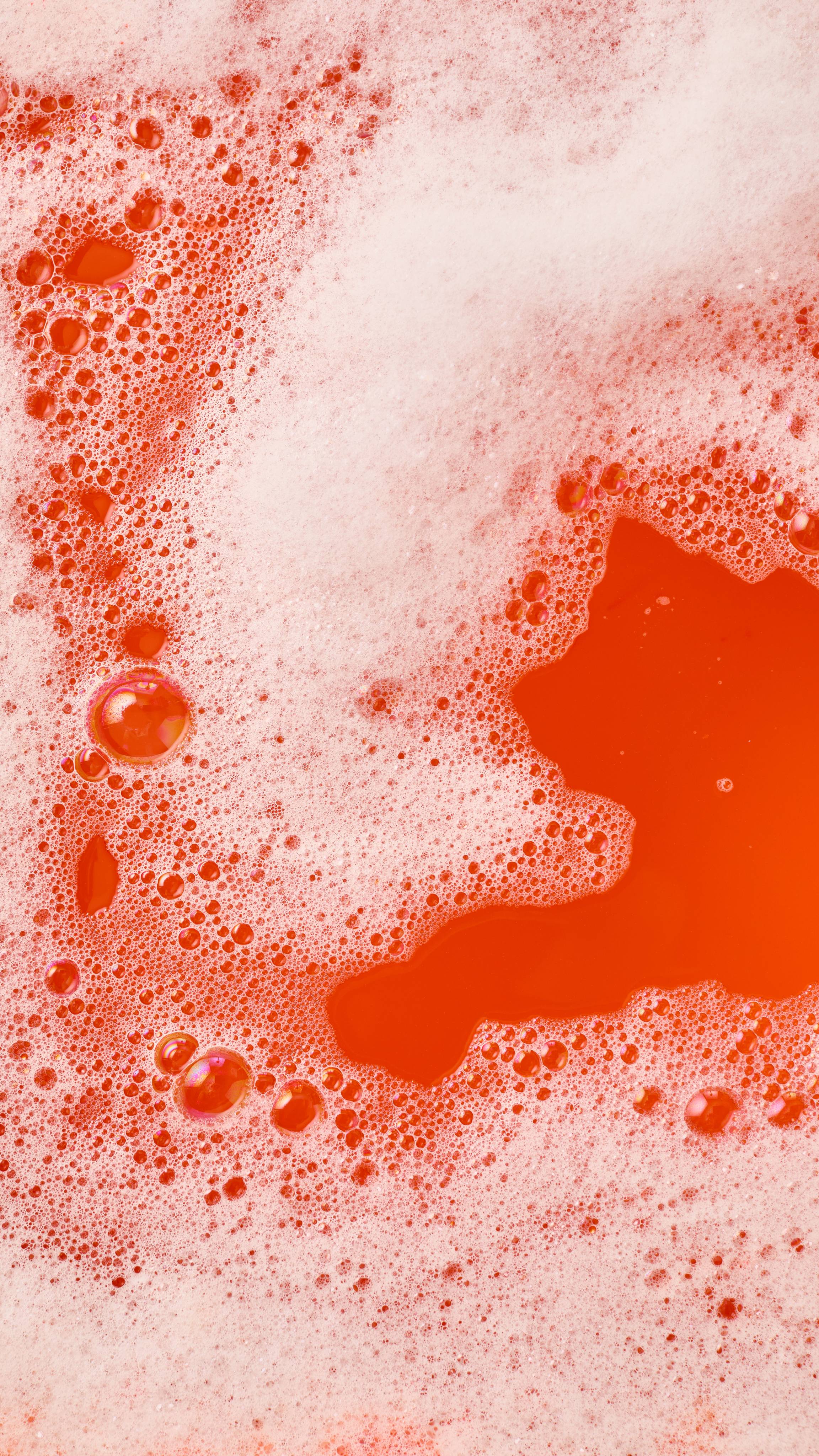 The image shows vibrant red-orange bath water largely covered in abundant white, frothy bubbles. The red bath water forms irregular shapes, winding through the foam, from the Toadstool-shaped 'Midnight Magic' bubble bar.