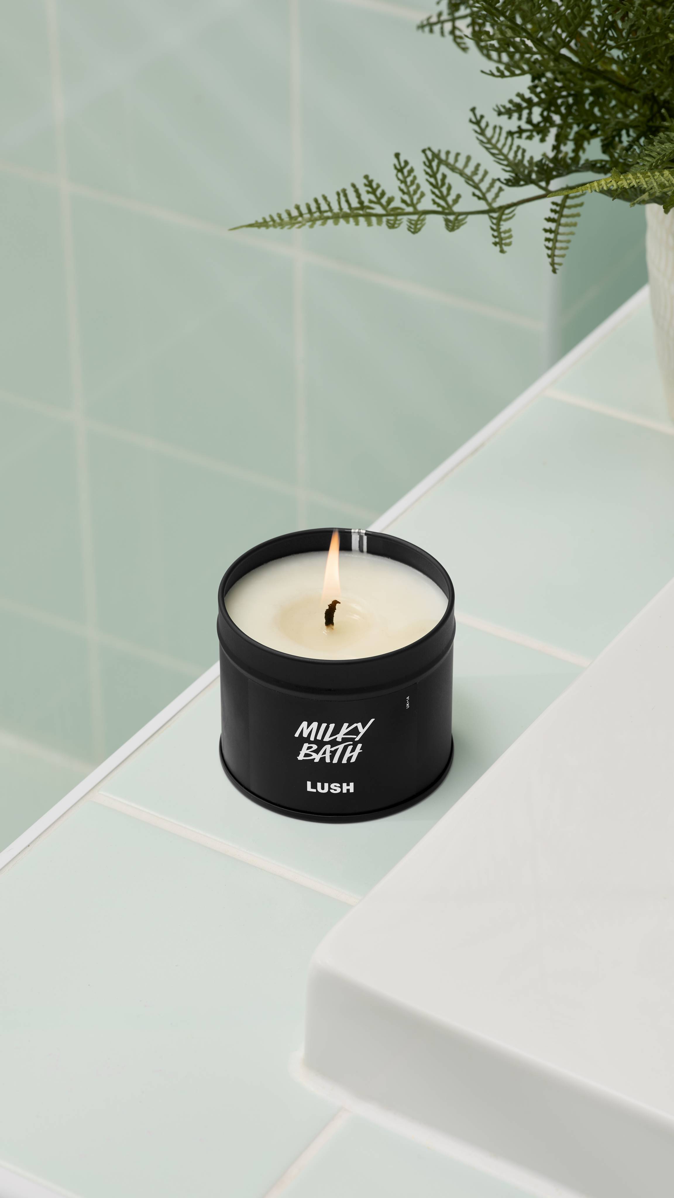 A close-up of an open, lit Milky Bath candle by Lush in a matte black tin, with the lid propped against the side. It sits on the edge of a white bathtub. Mint green tiles and a fern are in the background. All glitter used is plastic-free.