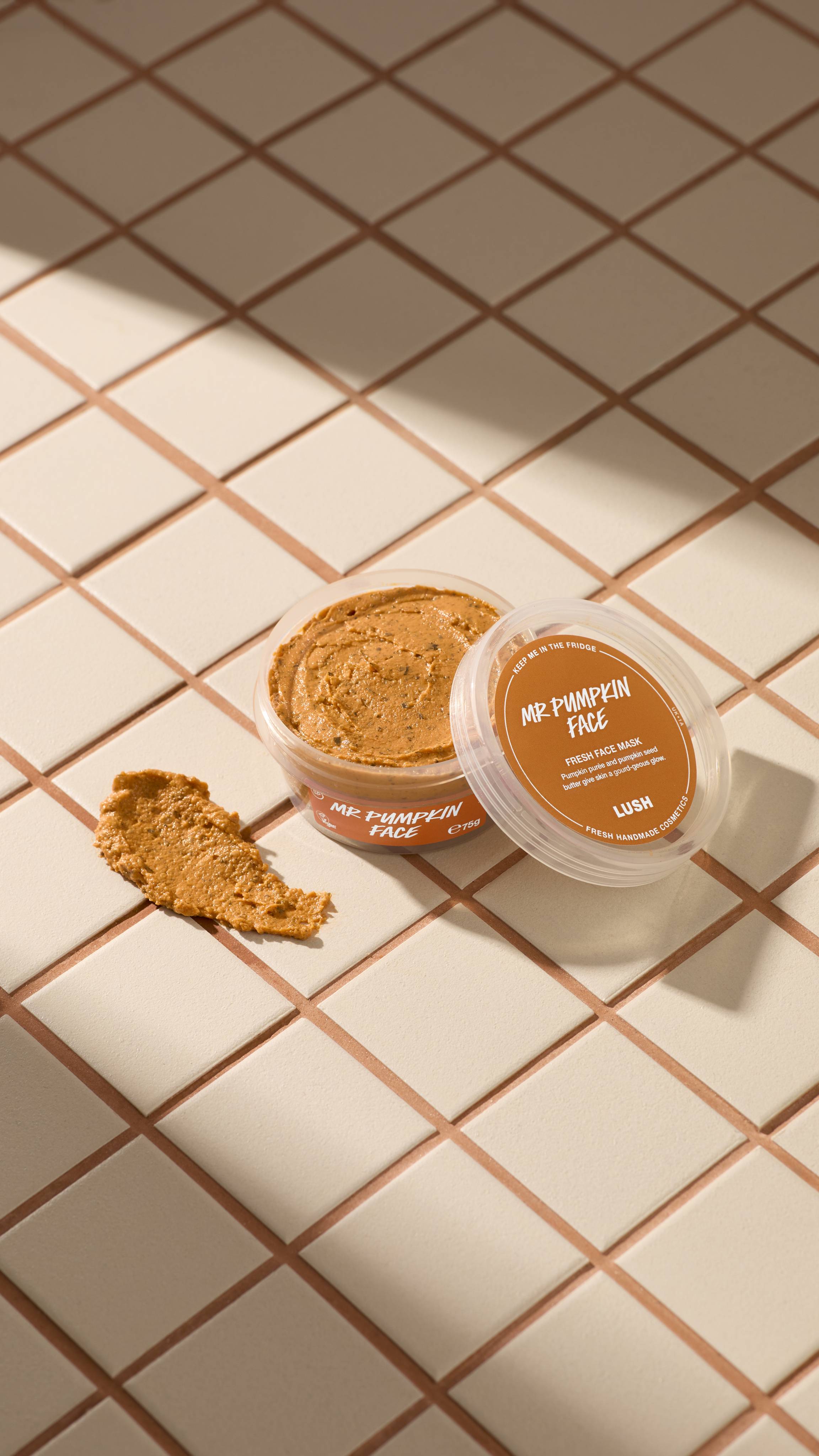 An open clear pot of Lush Mr Pumpkin Face fresh face mask sits on beige tiles next to a textured smear of the orange-brown mask. The clear lid with an orange label rests against the pot. Sunny shadows cross the tiled surface.
