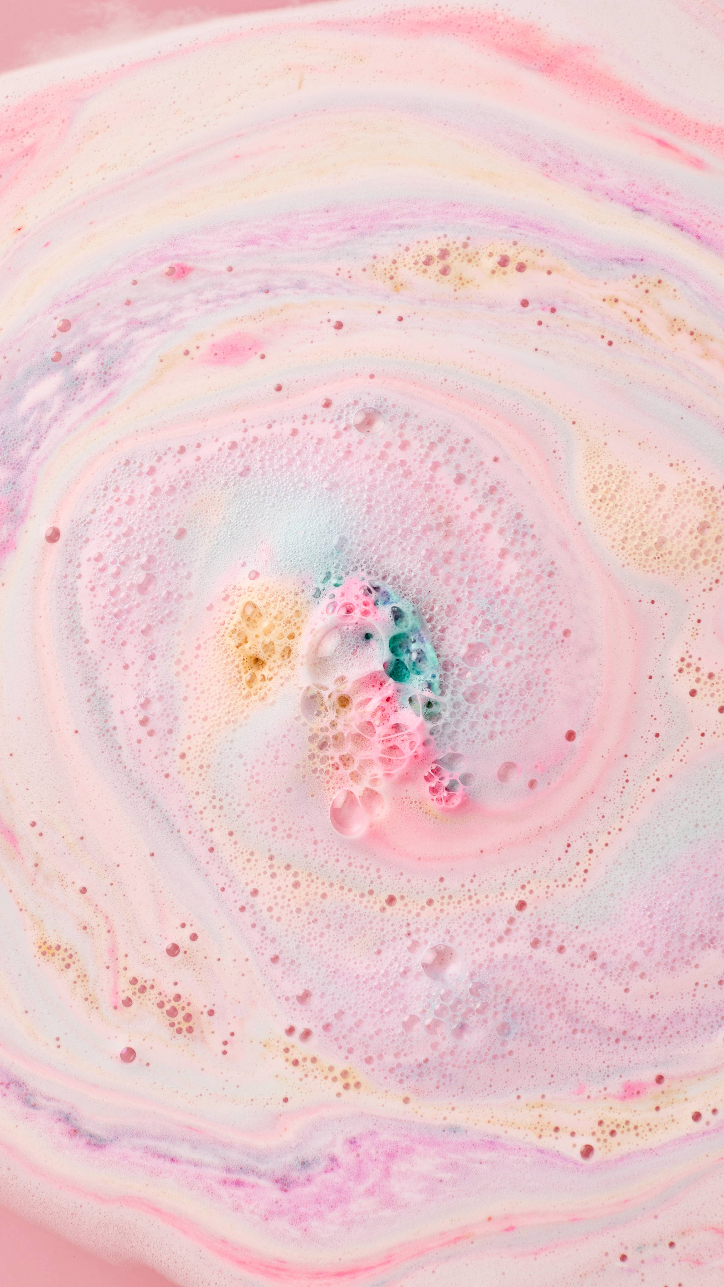 The image shows swirling bath art from the My Little Unicorn bath bomb, a dreamy blend of pastel pink, blue, yellow, and white hues. A colourful, bubbly core remains in the centre, fizzing and swirling the colours outwards.