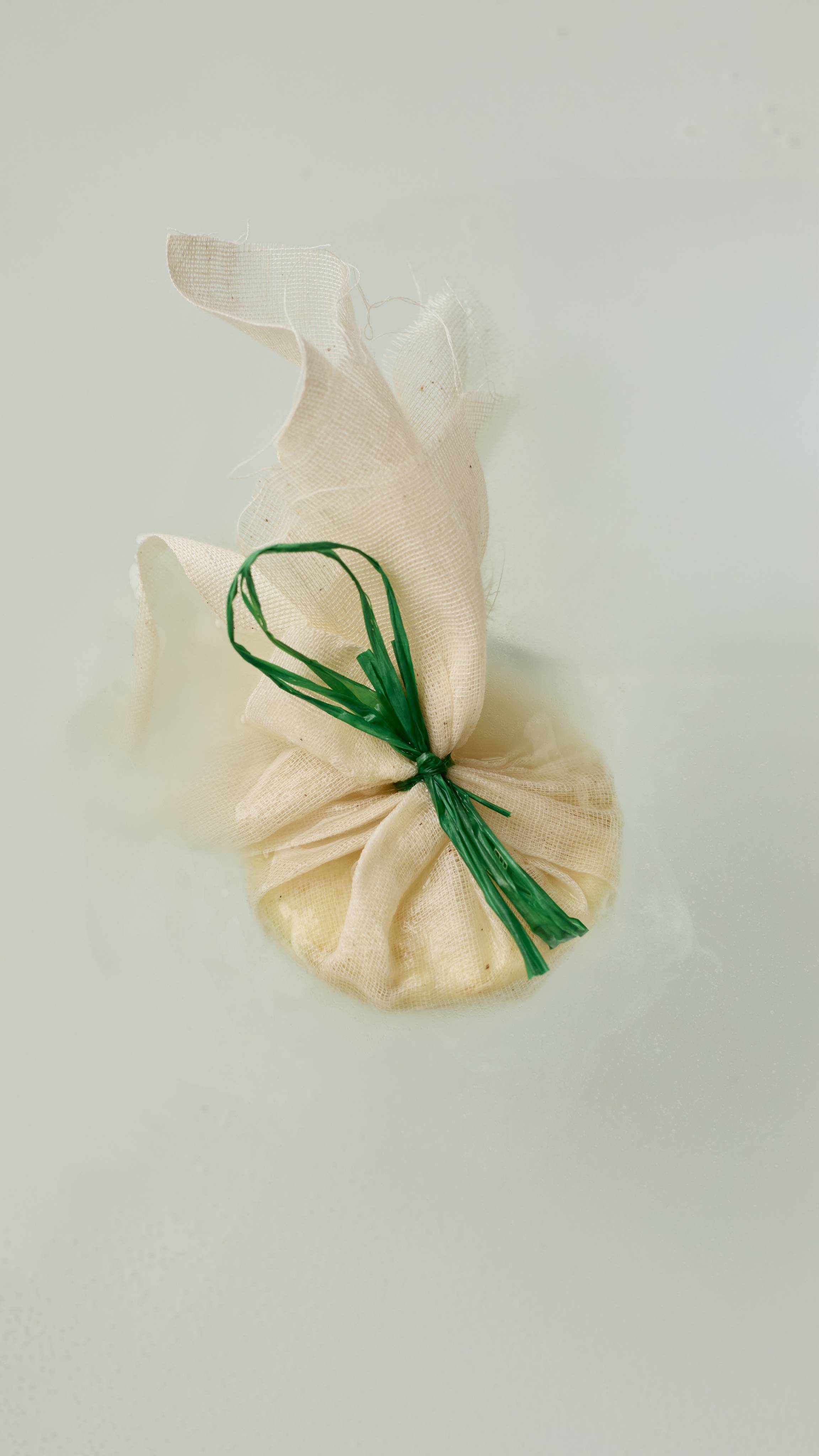 The image shows the Narcotick solid bath oil. A roughly textured muslin fabric pouch, gathered at the middle and tied with a vibrant green string. The top of the fabric flares out above the rounded product, which rests on clear water.