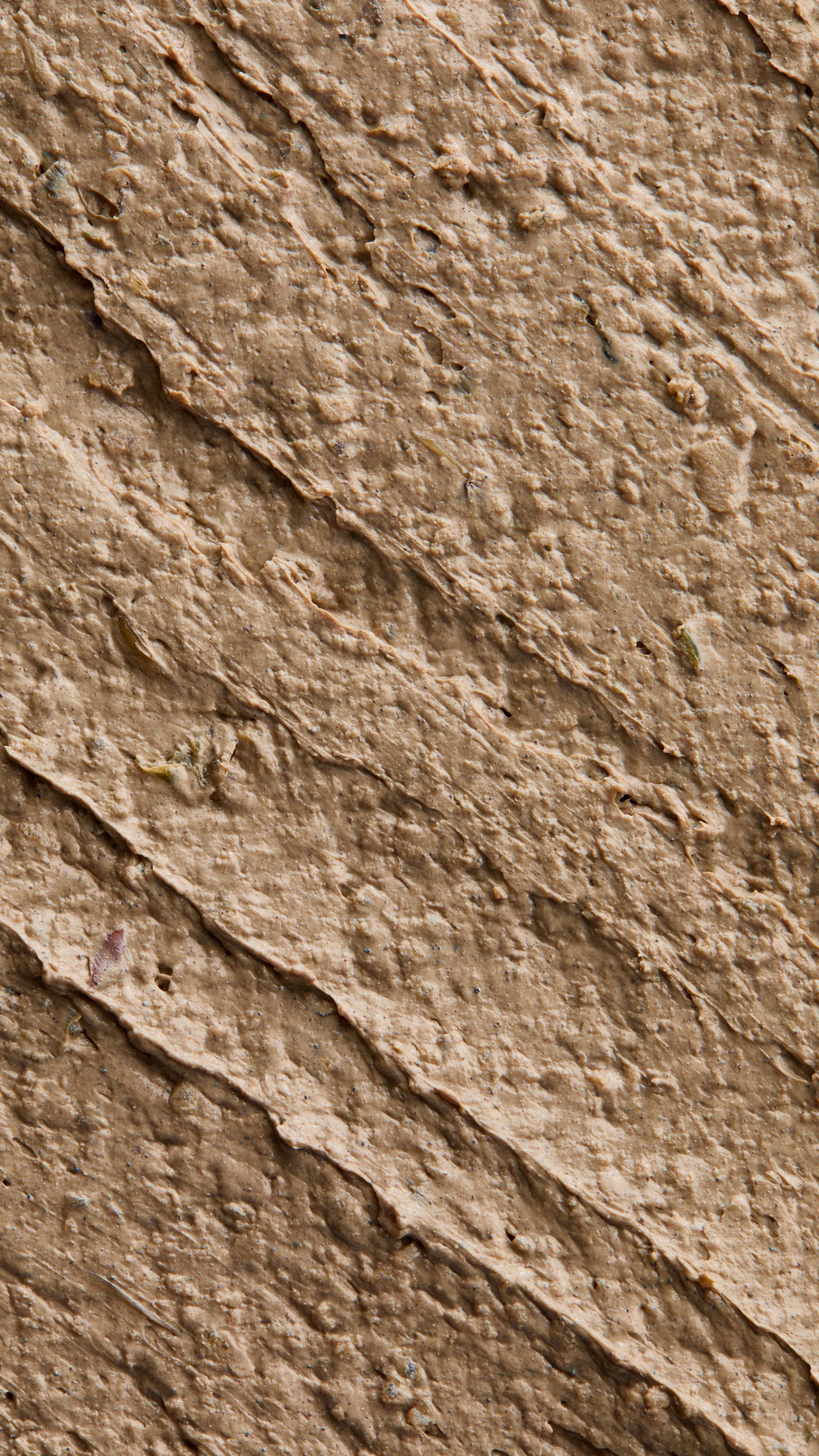 A close-up texture shot of thick, dark brown 'PADRON' foot mask, resembling rich soil. It features distinct, diagonal, rough streaks and visible flecks across its surface.
