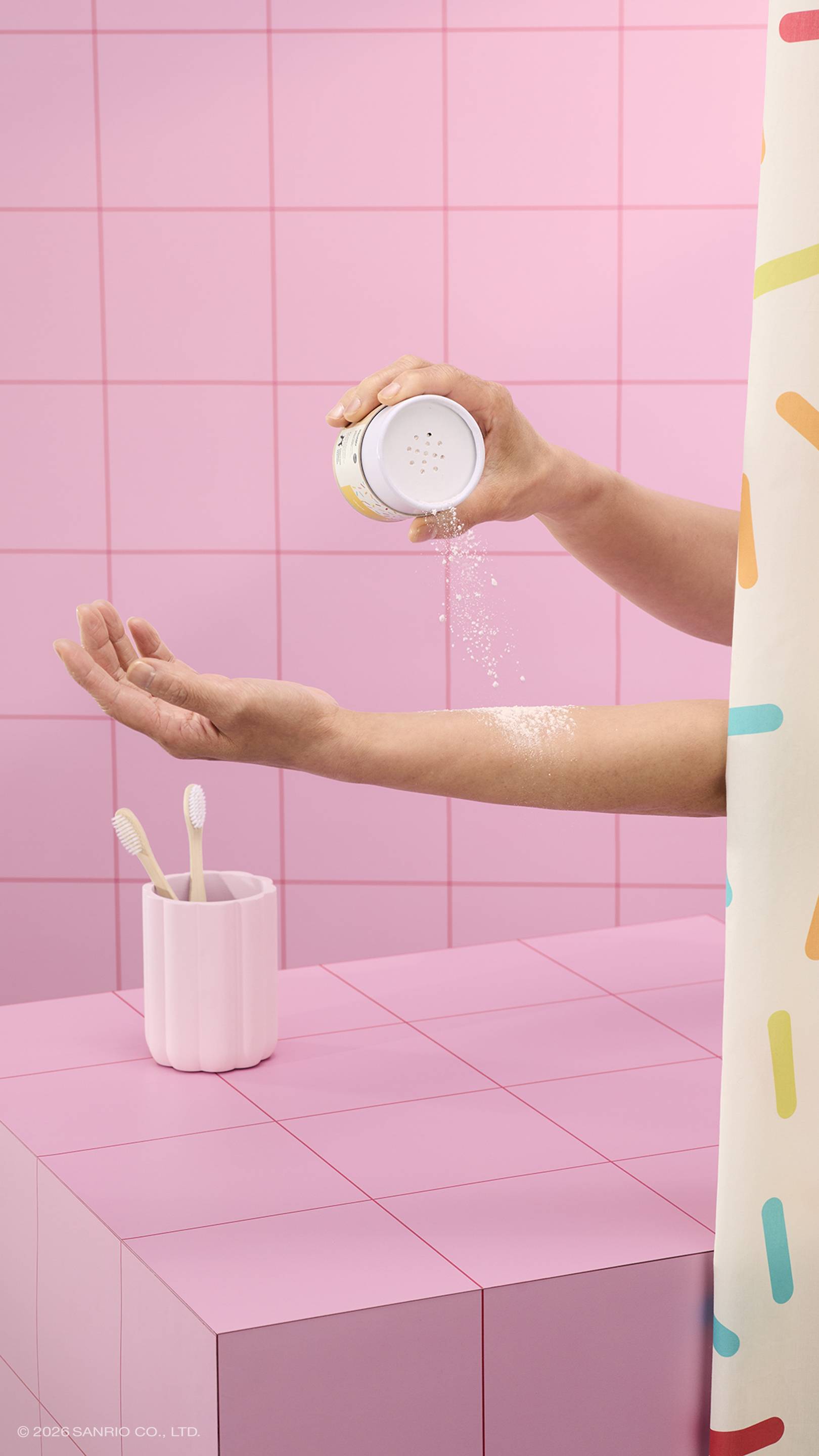 A hand shakes white dusting powder from a Lush x Pochacco tube onto another person's arm. The scene is set in a pink tiled bathroom with a sprinkle-patterned shower curtain and a toothbrush holder.