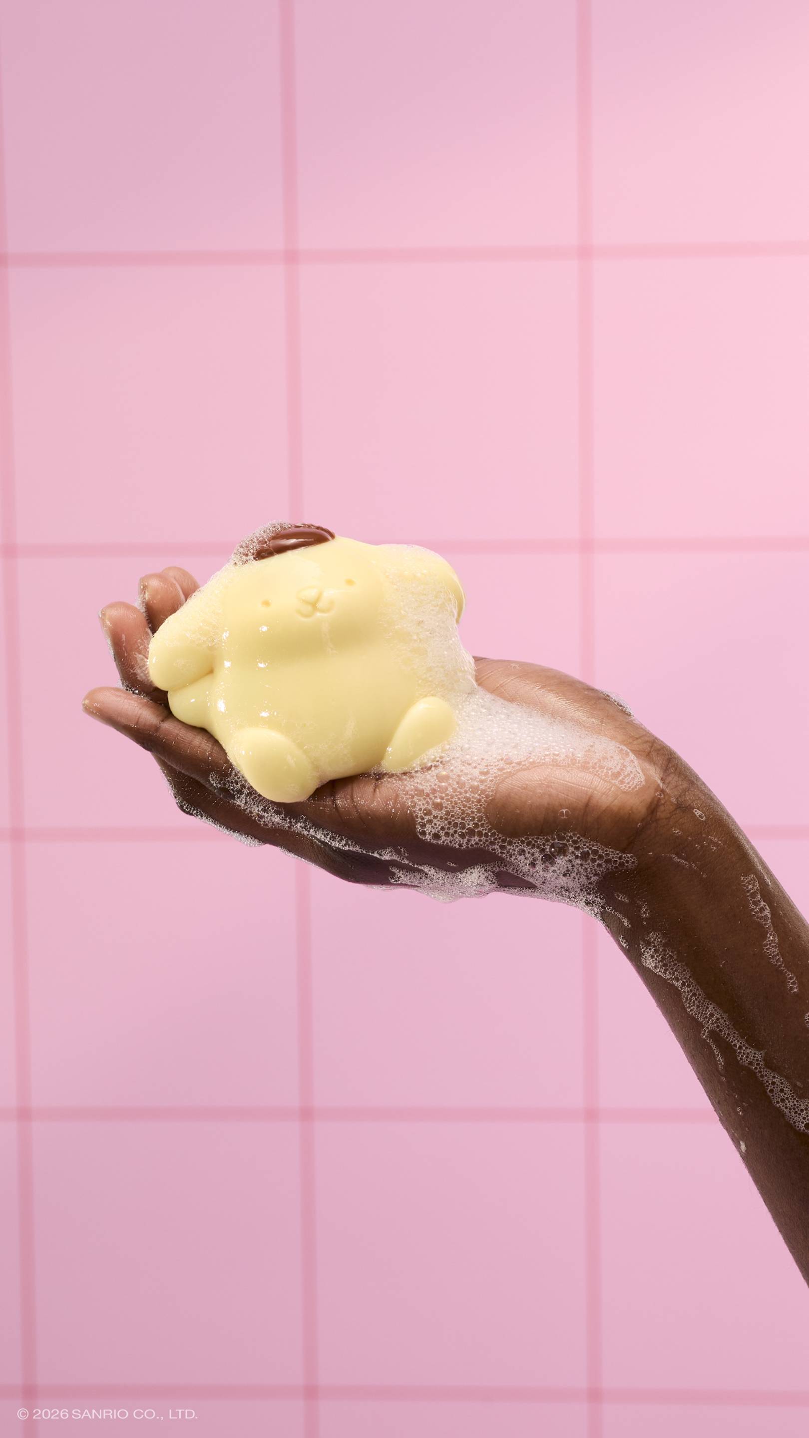 A soapy hand holds a glossy, pale yellow Pompompurin shower jelly, covered in white bubbles. The scene is set against a background of pink square tiles.