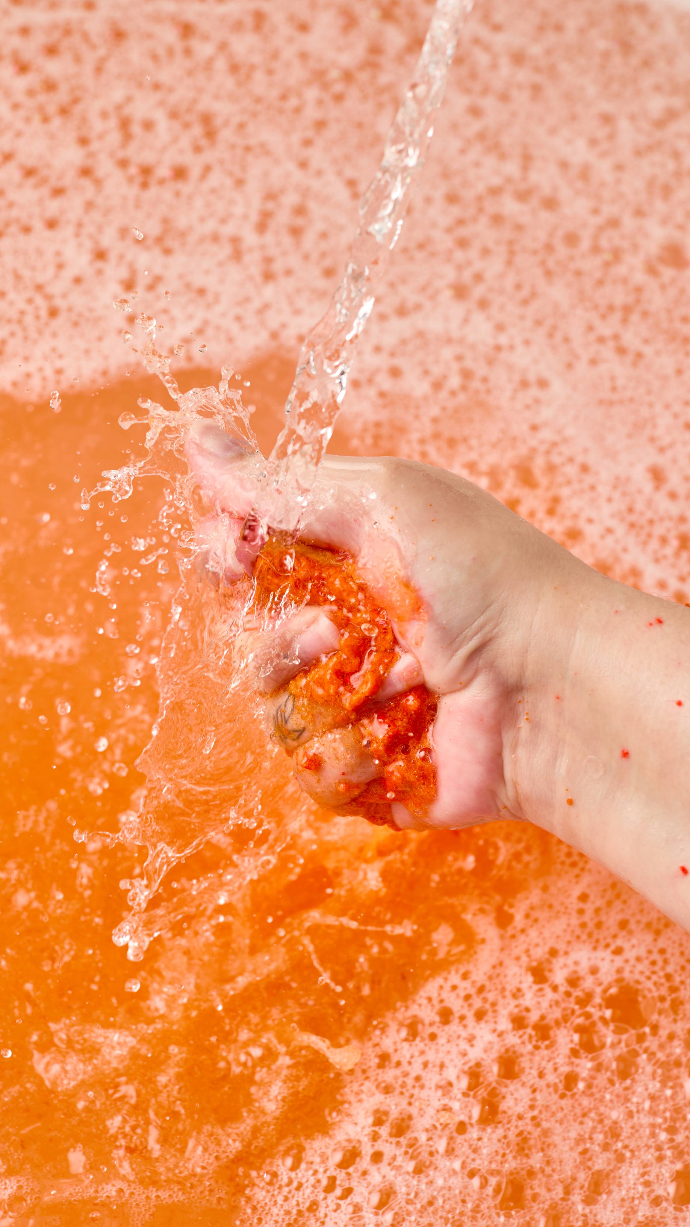 The image shows a light-toned hand vigorously squeezing and crumbling the gold and orange swirled 'Pumpkin Spice' bubble bar under a strong stream of water. Vibrant orange water is visible under a blanket of bubbles.