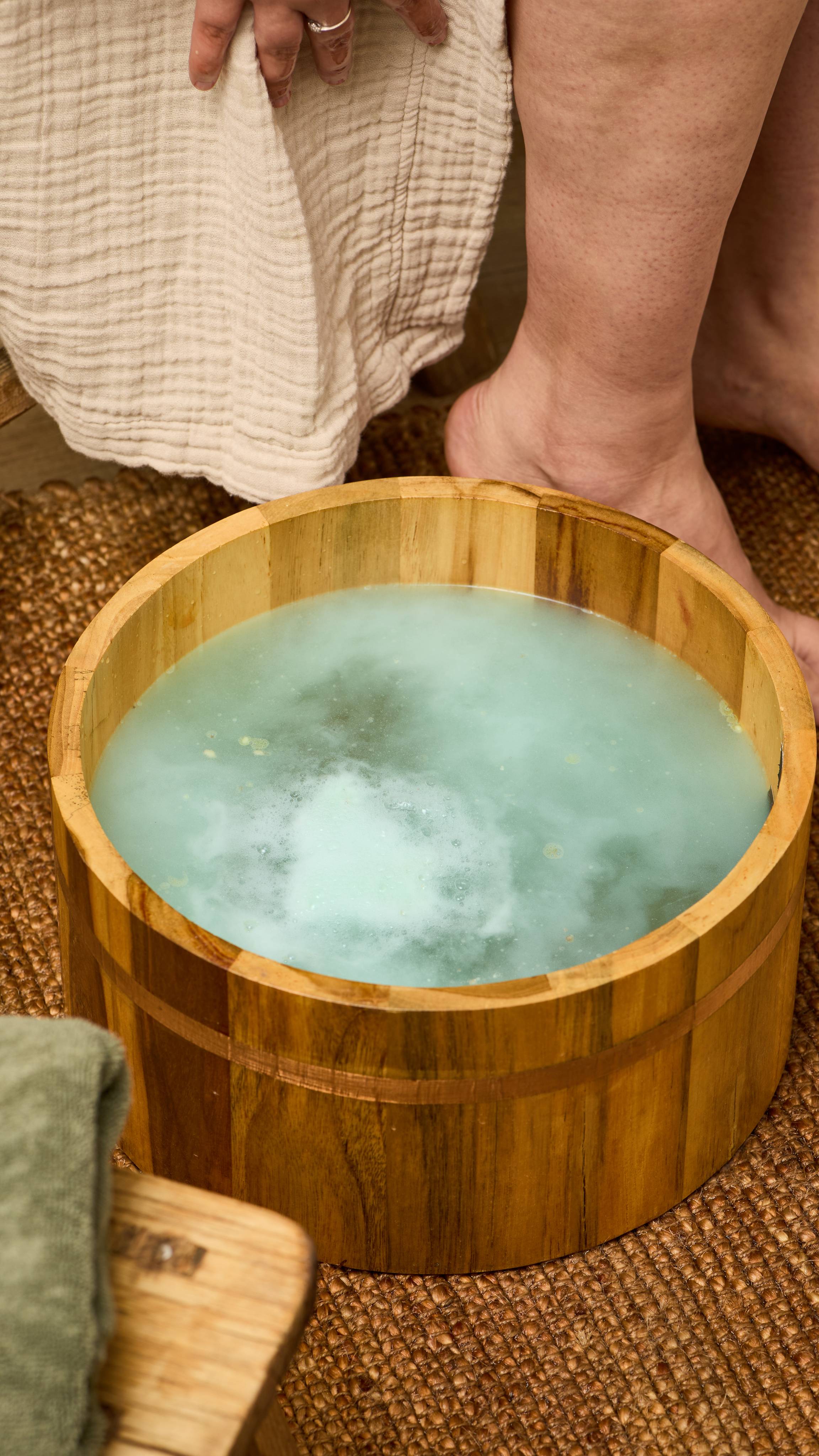 The image shows a wooden foot bath tub filled with light green water, creating a foamy surface. It rests on a woven mat, with a light-coloured towel draped nearby and blurred bare legs in the background.